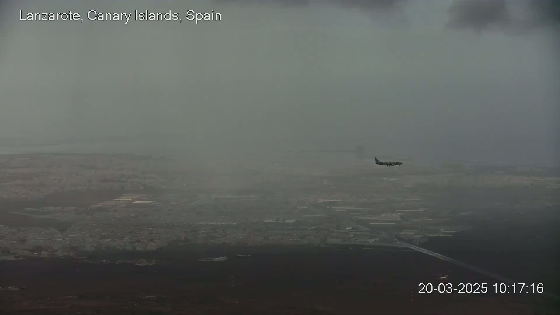 A plane flies over a hazy, overcast city and ocean.