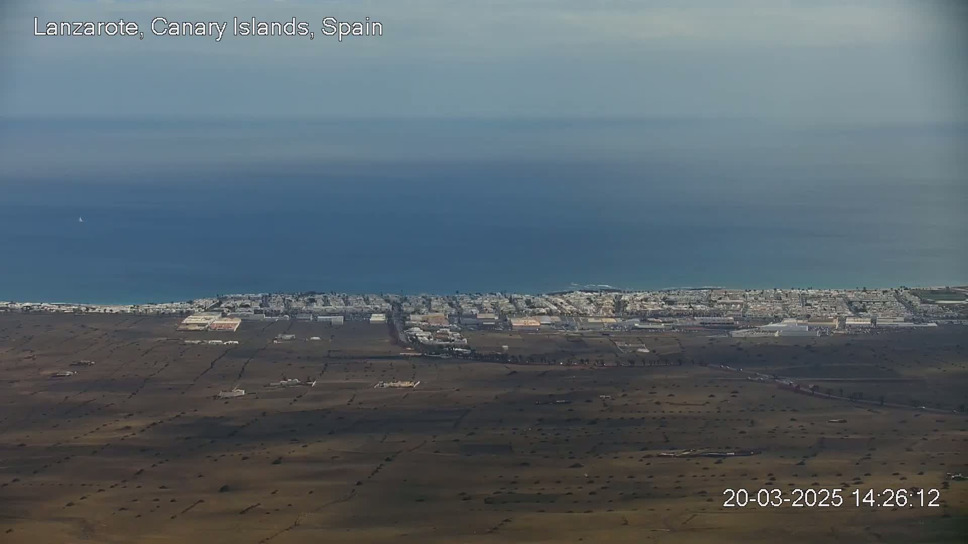 An aerial view shows a coastal town situated on a mostly barren, dry landscape next to a calm, blue ocean under a clear sky.