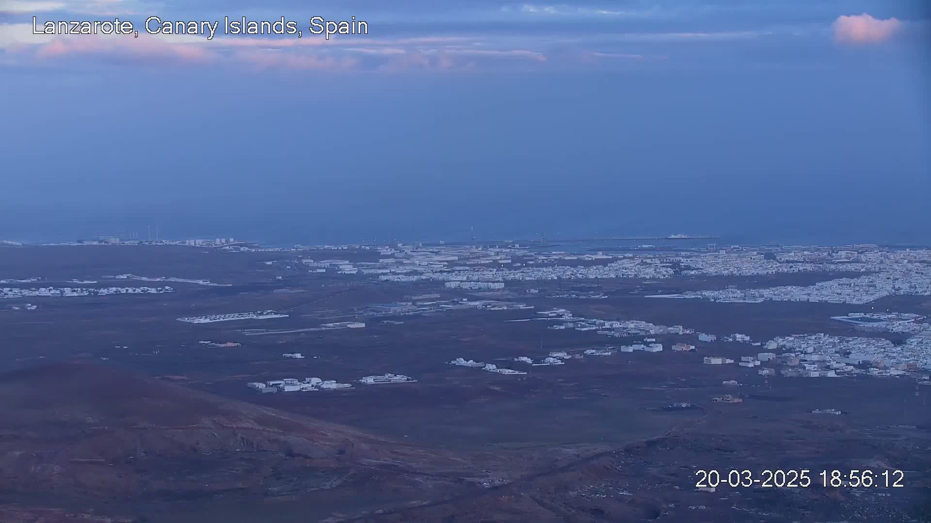 A sprawling coastal town with white buildings stretches across a dark volcanic landscape under a twilight sky.