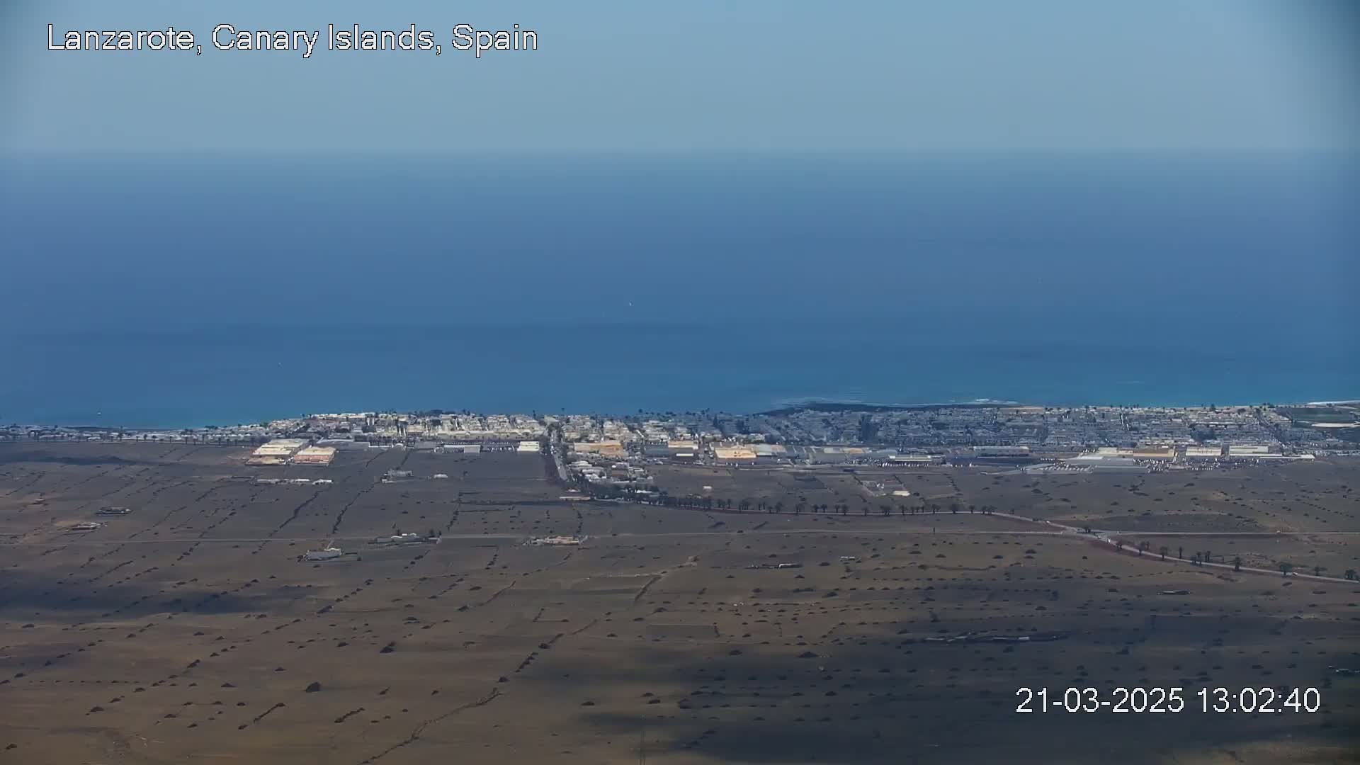 A high-angle view shows a coastal town on a largely flat, arid landscape next to a calm ocean under a clear sky.