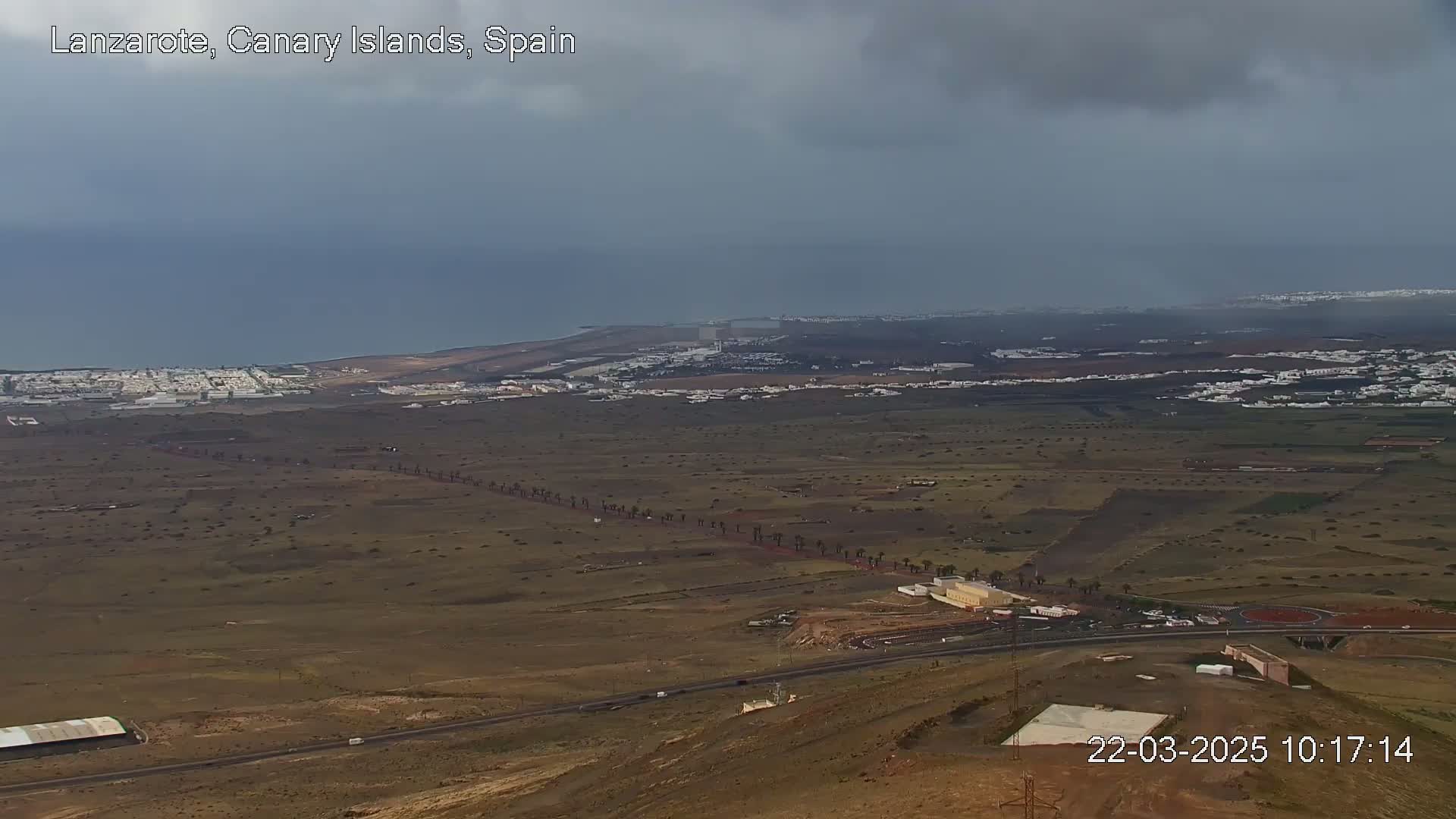 A wide, high-angle view shows a dry, brown landscape with a coastal town in the distance under a cloudy sky.