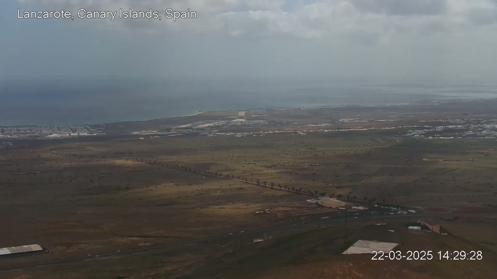 A high-angle, long shot shows a mostly-flat, brown and sparsely vegetated landscape with a coastal town and ocean in the distance under a mostly cloudy sky.