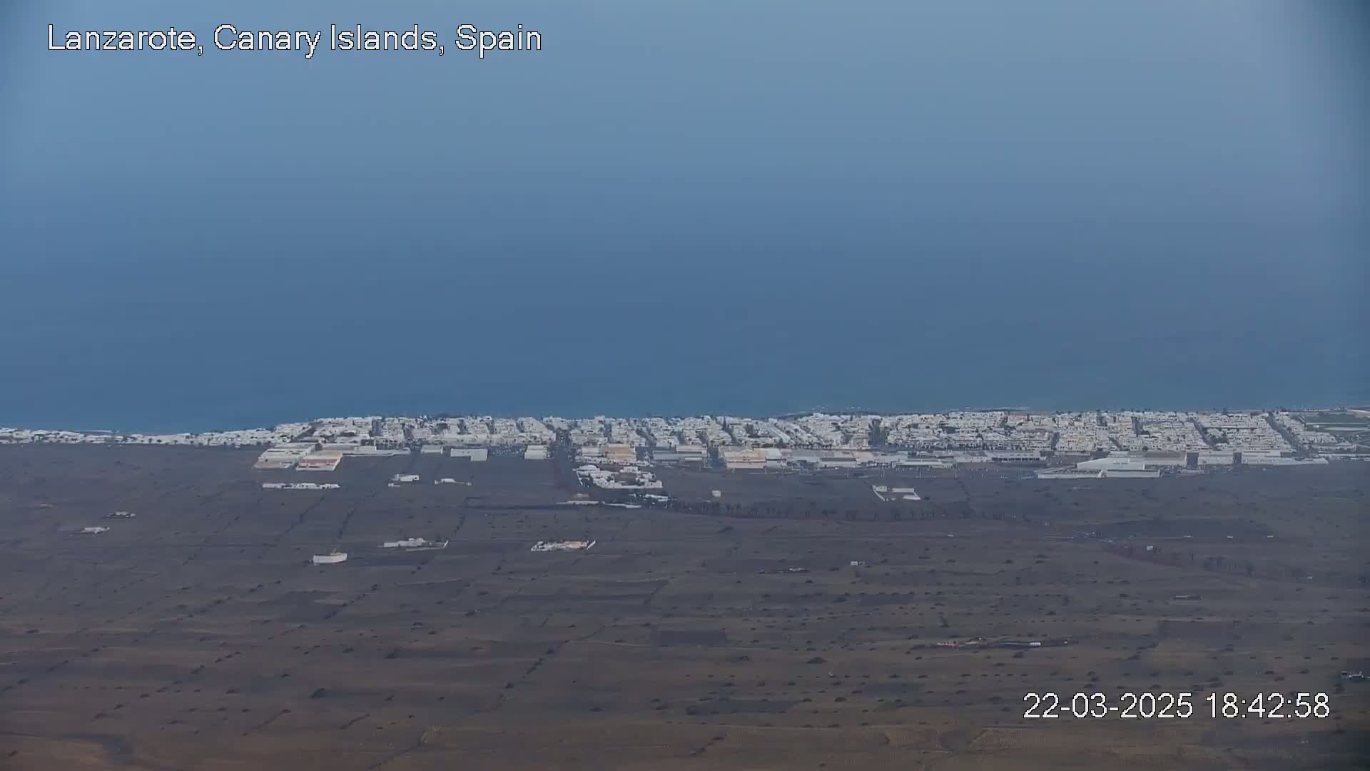 A coastal town with white buildings stretches along a dark-colored, sparsely vegetated plain, under a clear, pale blue sky next to a calm ocean.