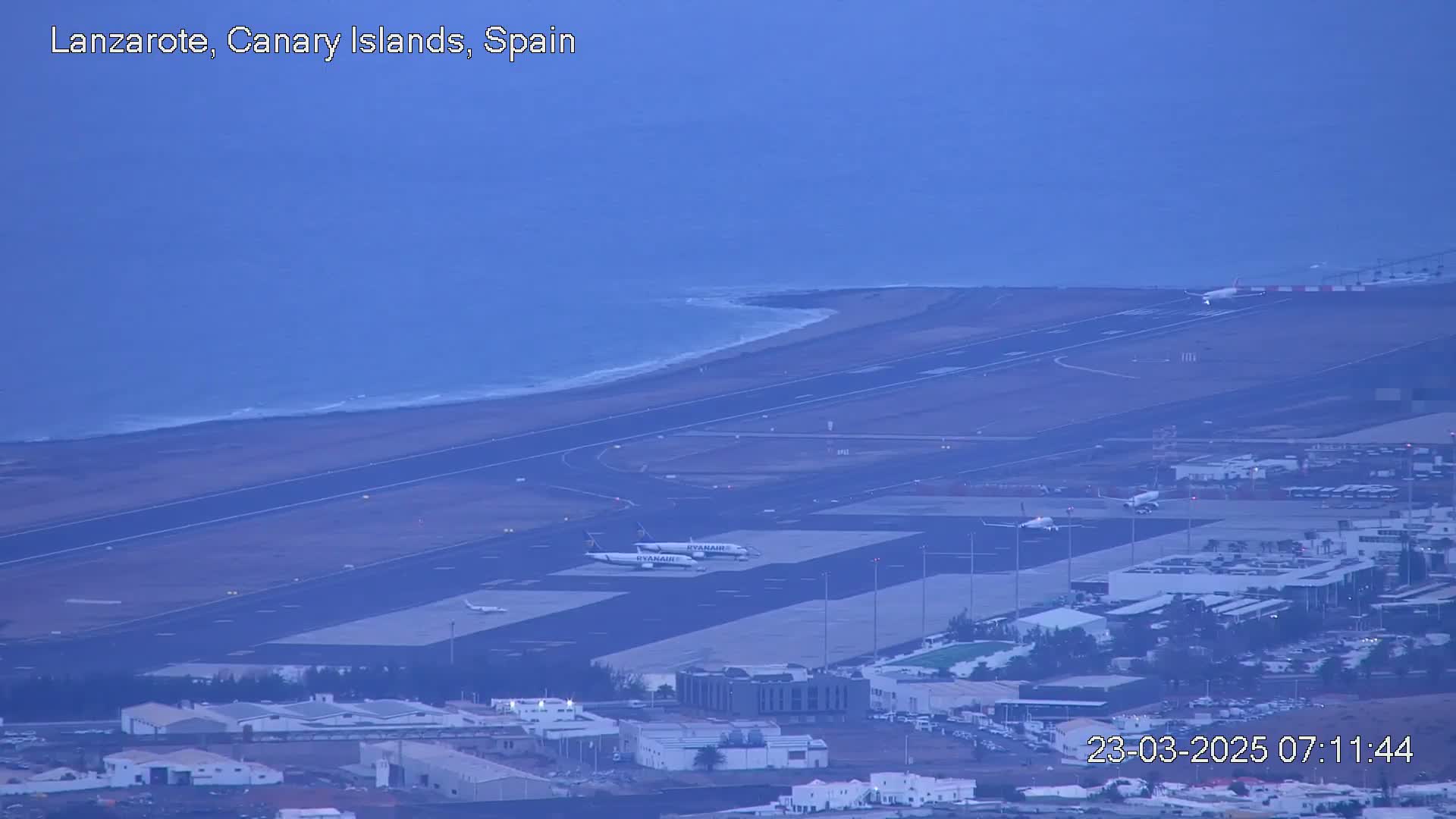 An airport with several airplanes on the tarmac and a coastline with ocean waves visible in the distance under a hazy sky.