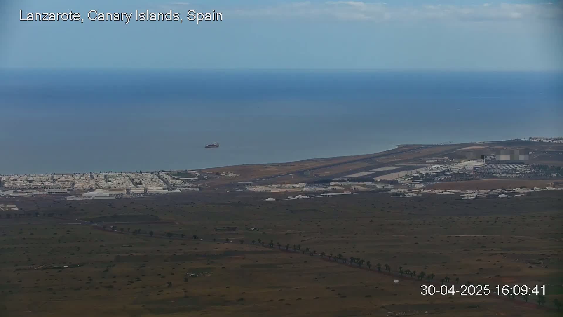 A high-angle, long shot shows a coastal town, an airport, and a calm ocean under a mostly clear sky.