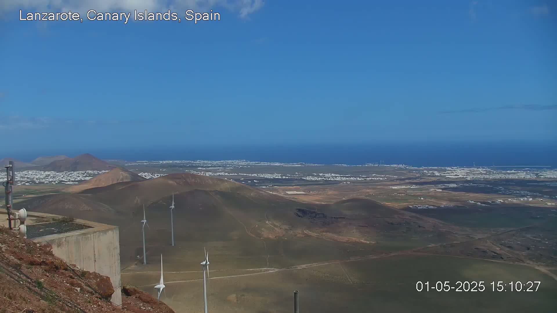 A panoramic view on a clear day shows a coastal town nestled among volcanic hills, with wind turbines scattered across the landscape and a calm ocean in the distance.