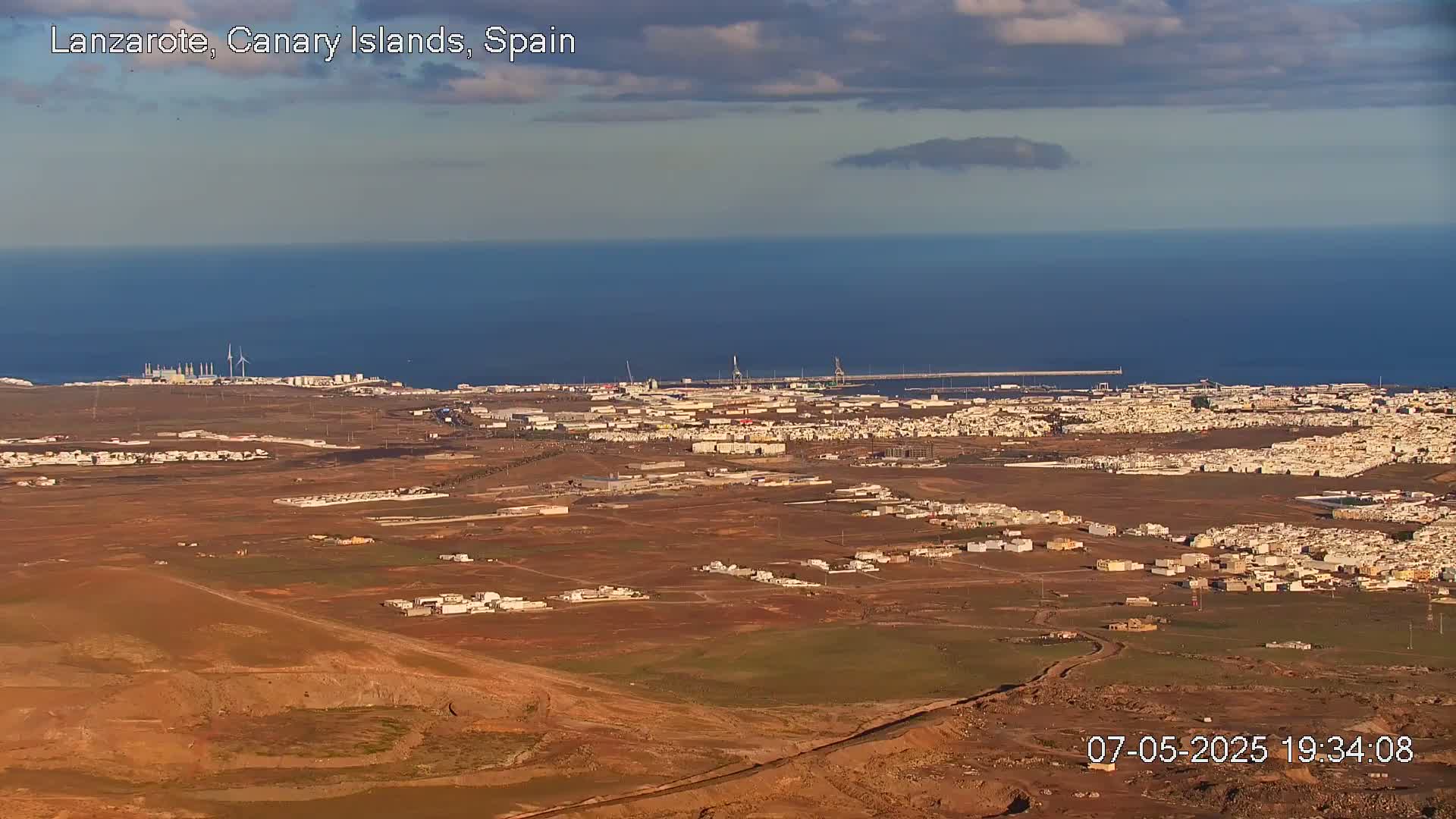 A sprawling coastal town with low-rise buildings extends across a dry, reddish-brown landscape under a partly cloudy sky, overlooking a calm ocean.