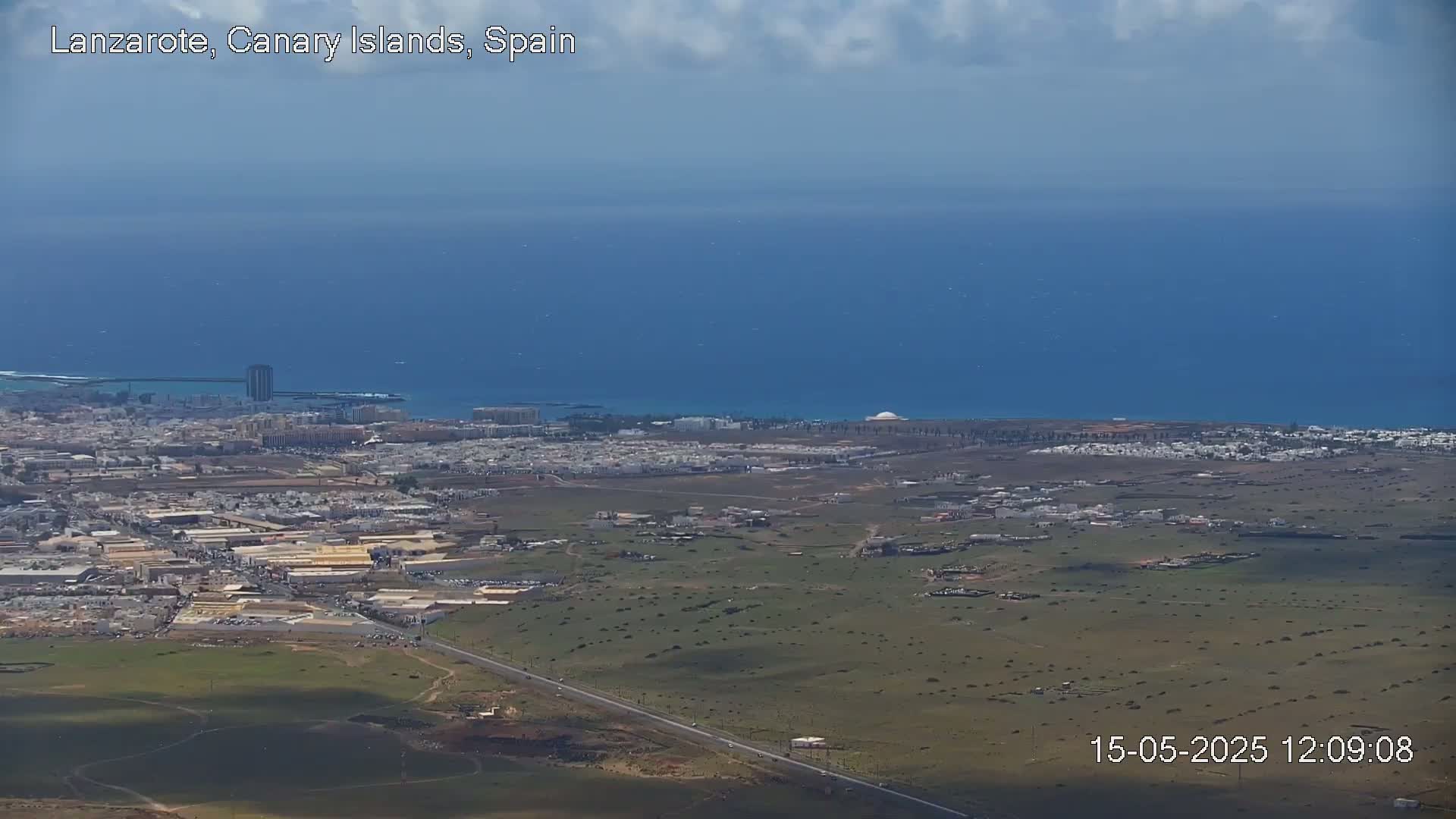 A coastal city with low-rise buildings sprawls across a dry, arid landscape, meeting a calm, blue ocean under a partly cloudy sky.