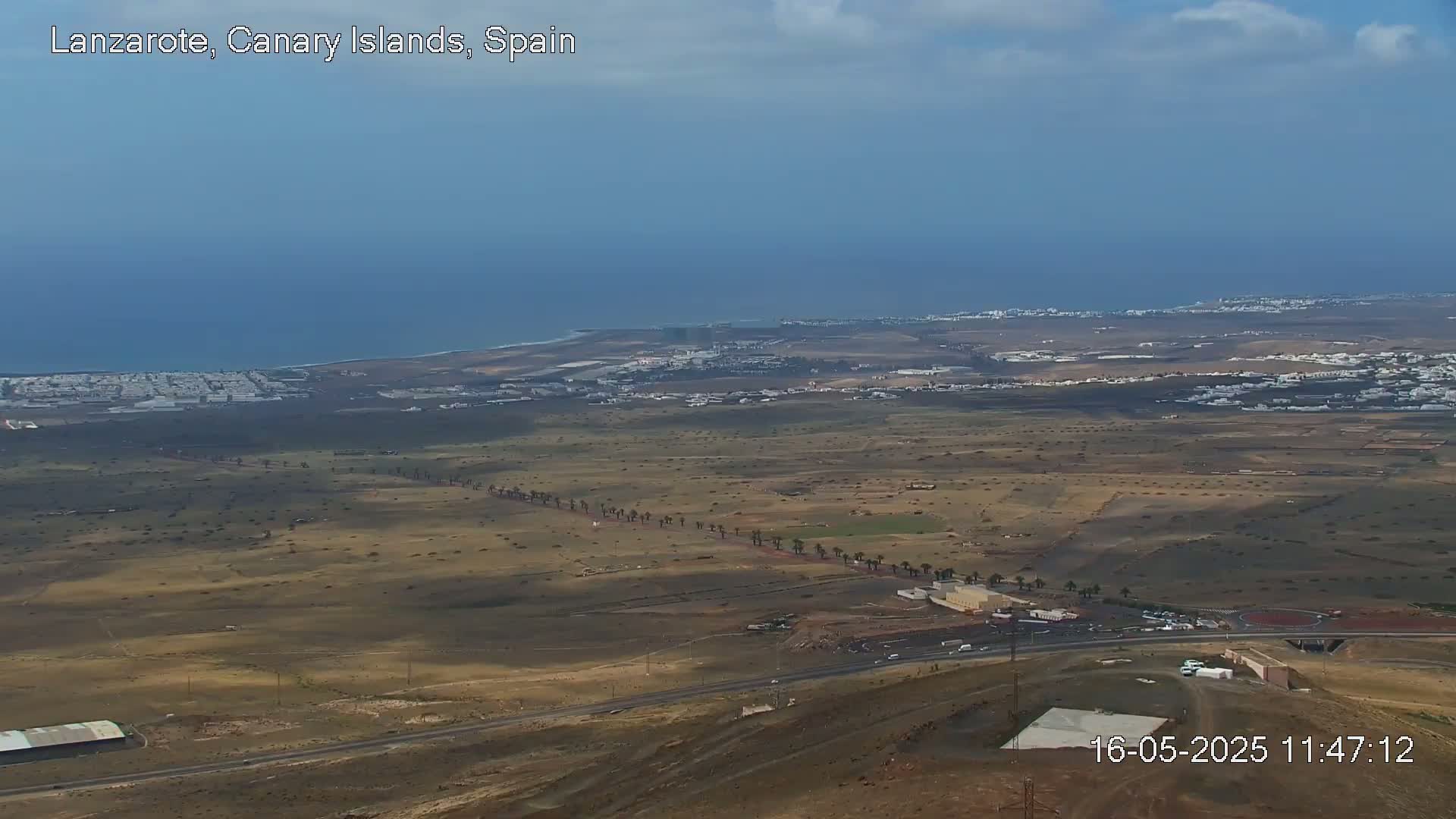 A high-angle, long shot shows a mostly sunny, arid landscape with a coastal town and ocean in the distance under a partly cloudy sky.