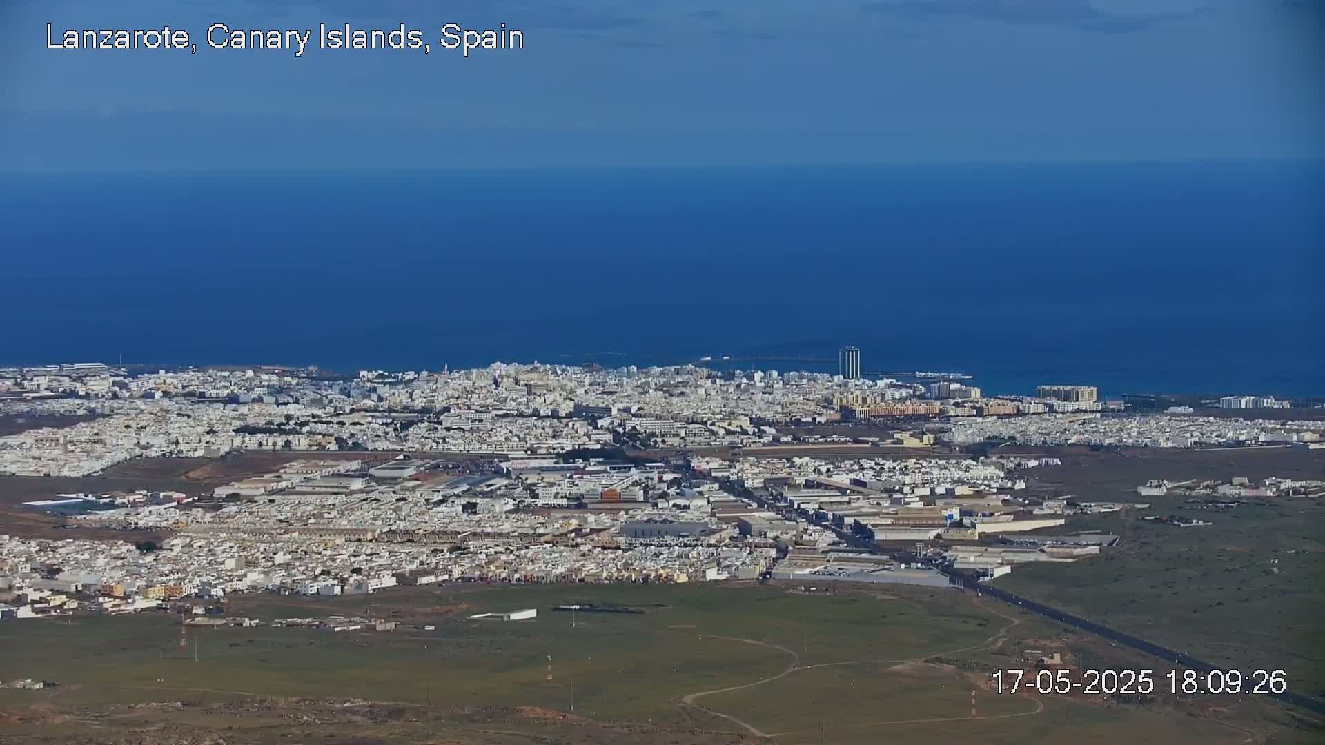 An aerial view shows a coastal city with low-rise buildings under a clear blue sky and calm blue ocean.