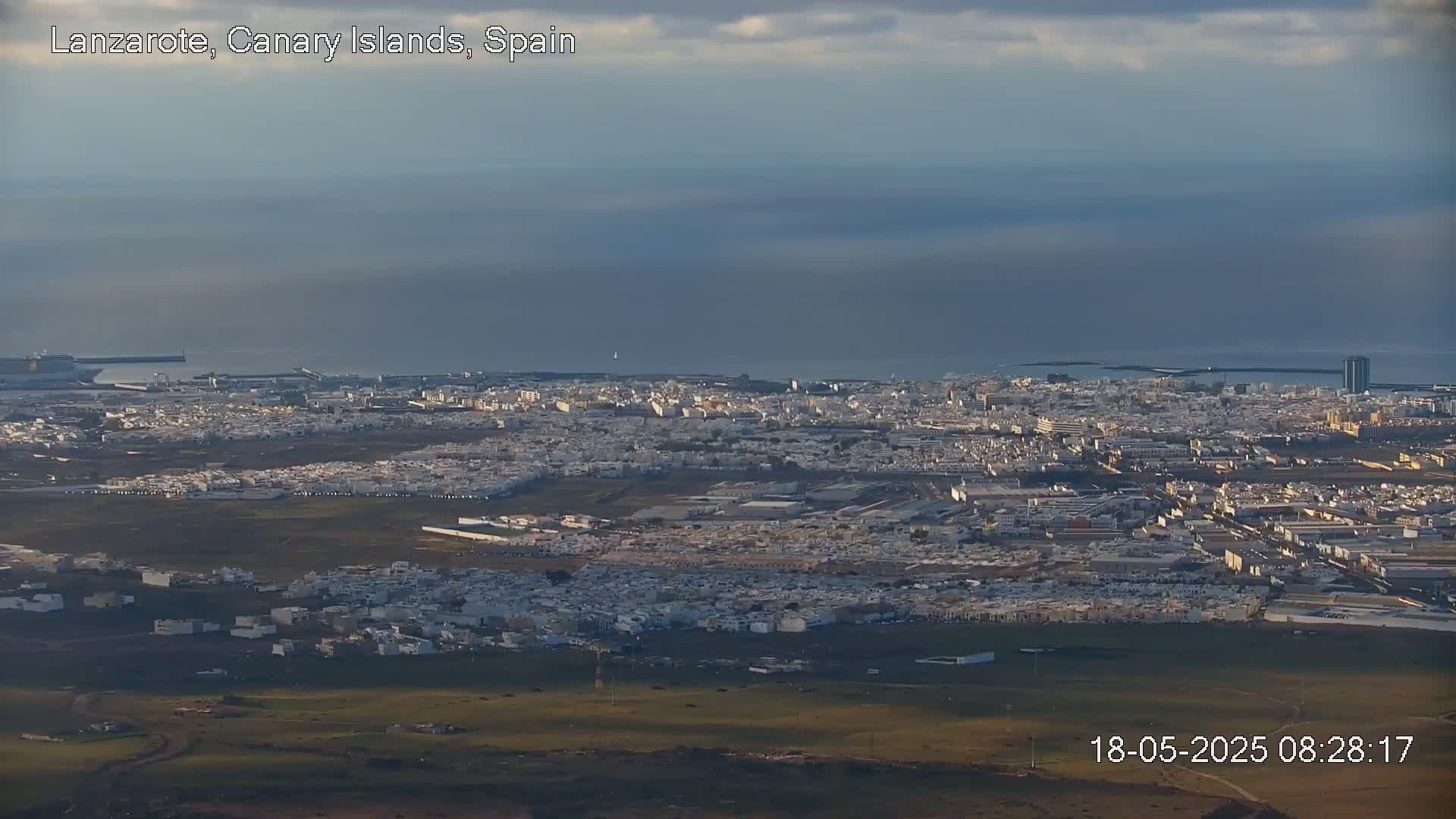 An expansive view of a coastal city under a mostly cloudy sky shows a large expanse of low white buildings extending to a harbor and ocean.