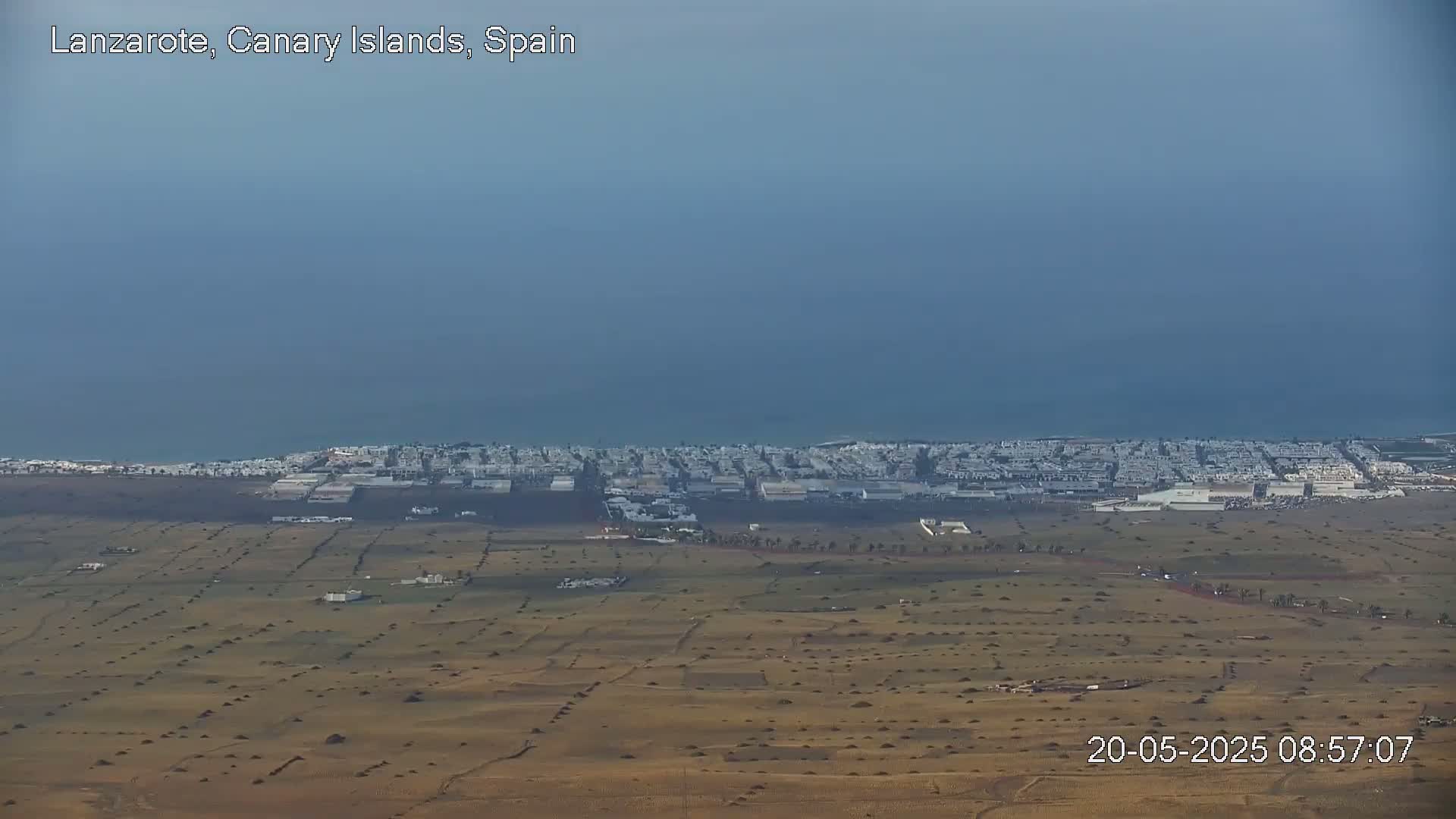 A coastal town with white buildings stretches along a hazy, blue ocean under a mostly clear sky.