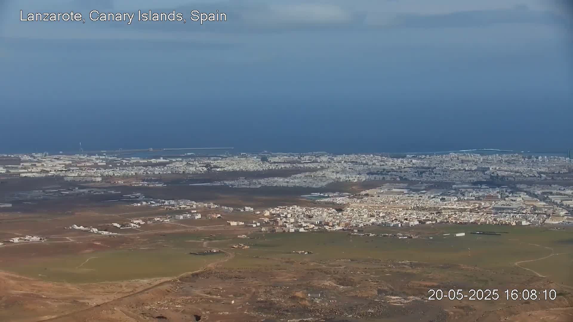 A sprawling coastal city with light-brown and tan buildings extends to a dark-blue ocean under a mostly clear sky.
