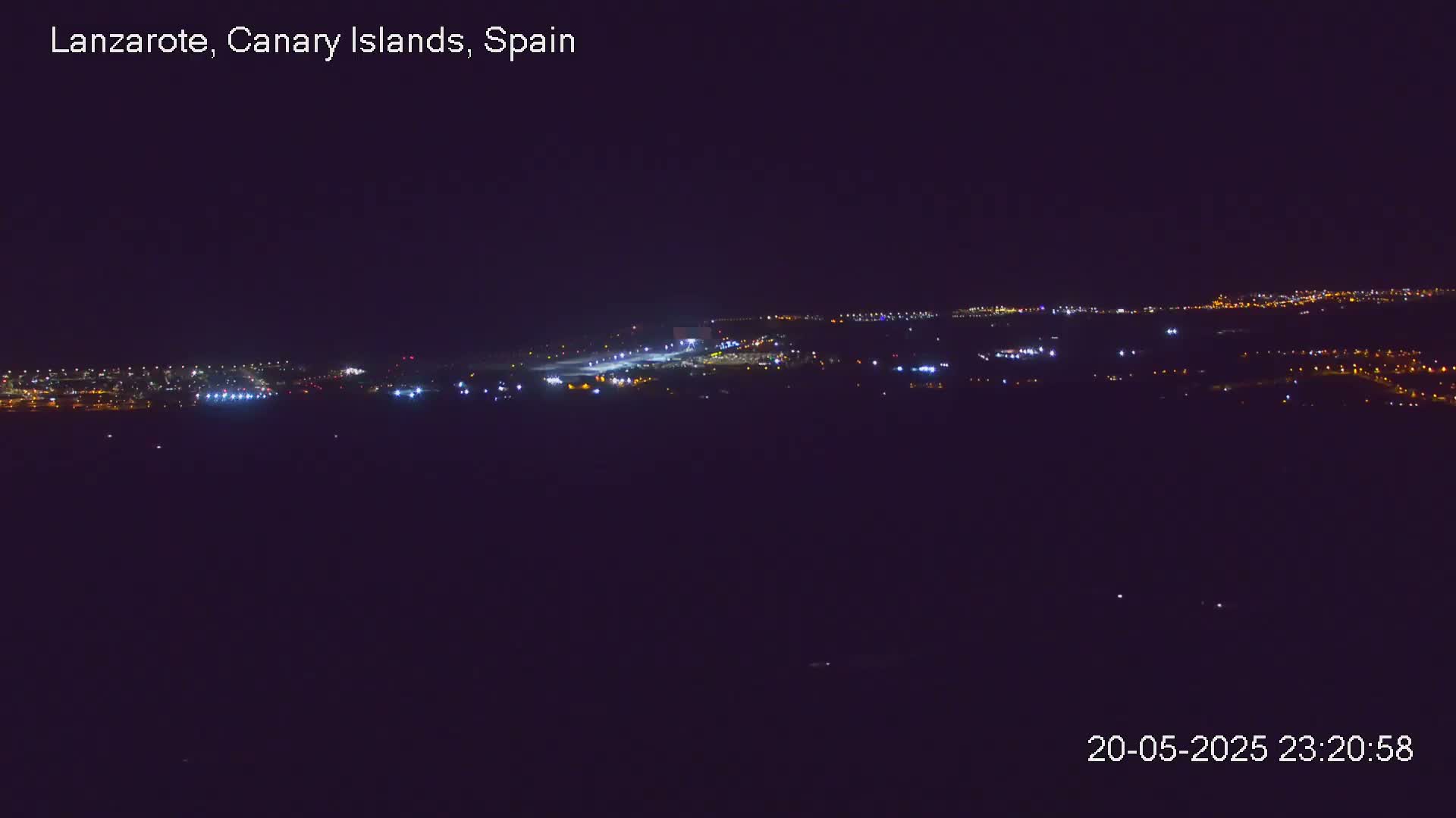 A nighttime long shot shows an airport runway and surrounding buildings illuminated, under a clear dark sky.