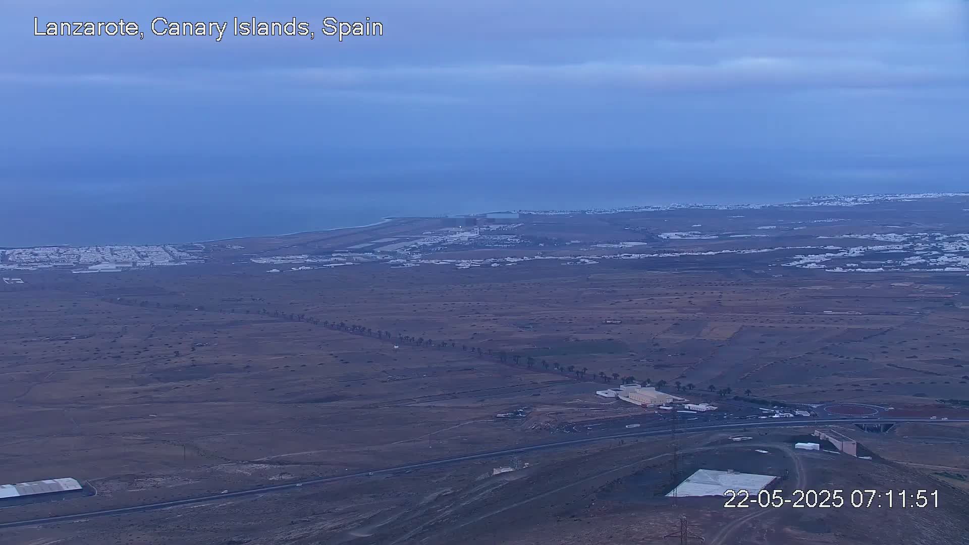 A high-angle, long shot shows a coastal town and surrounding arid landscape under a mostly clear, dusky blue sky.