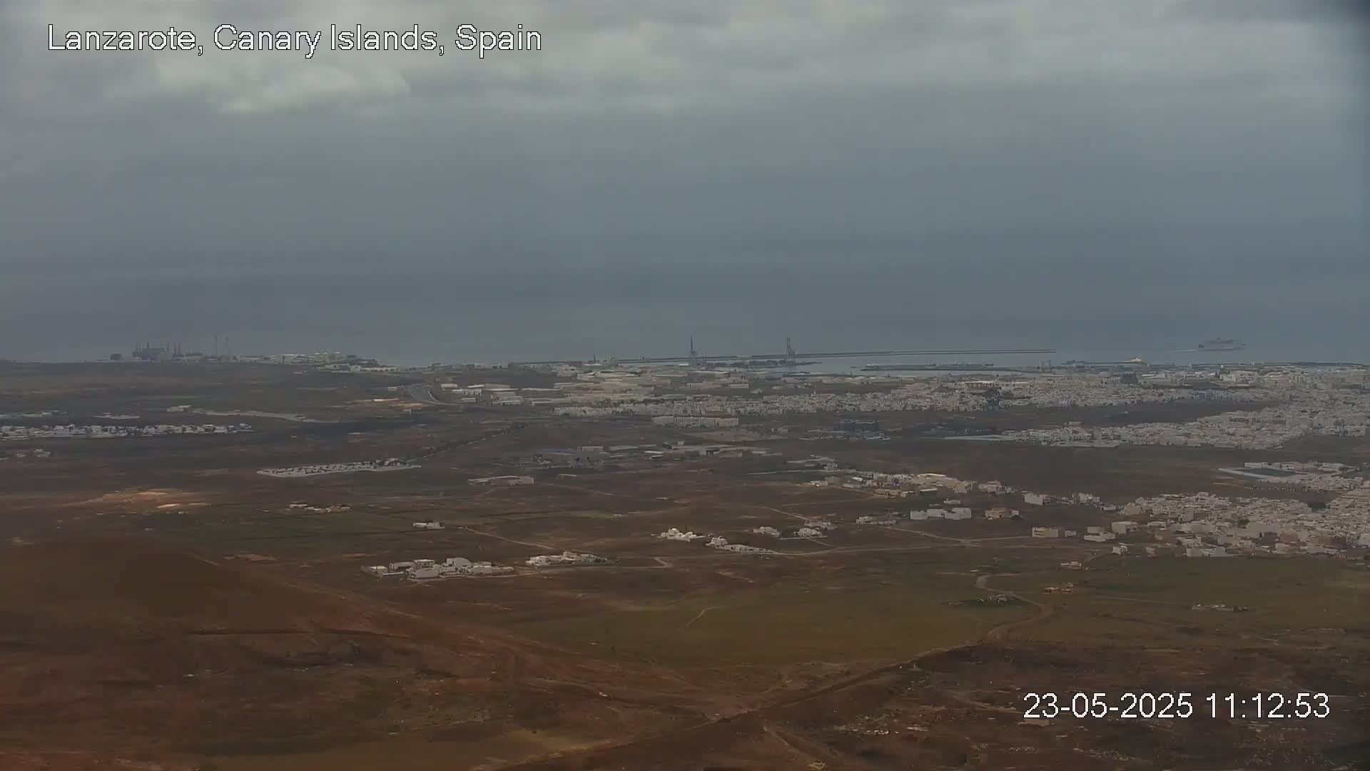 A high-angle view of a coastal town under an overcast sky shows a sprawling landscape of low buildings and brown earth extending to a distant port and ocean.
