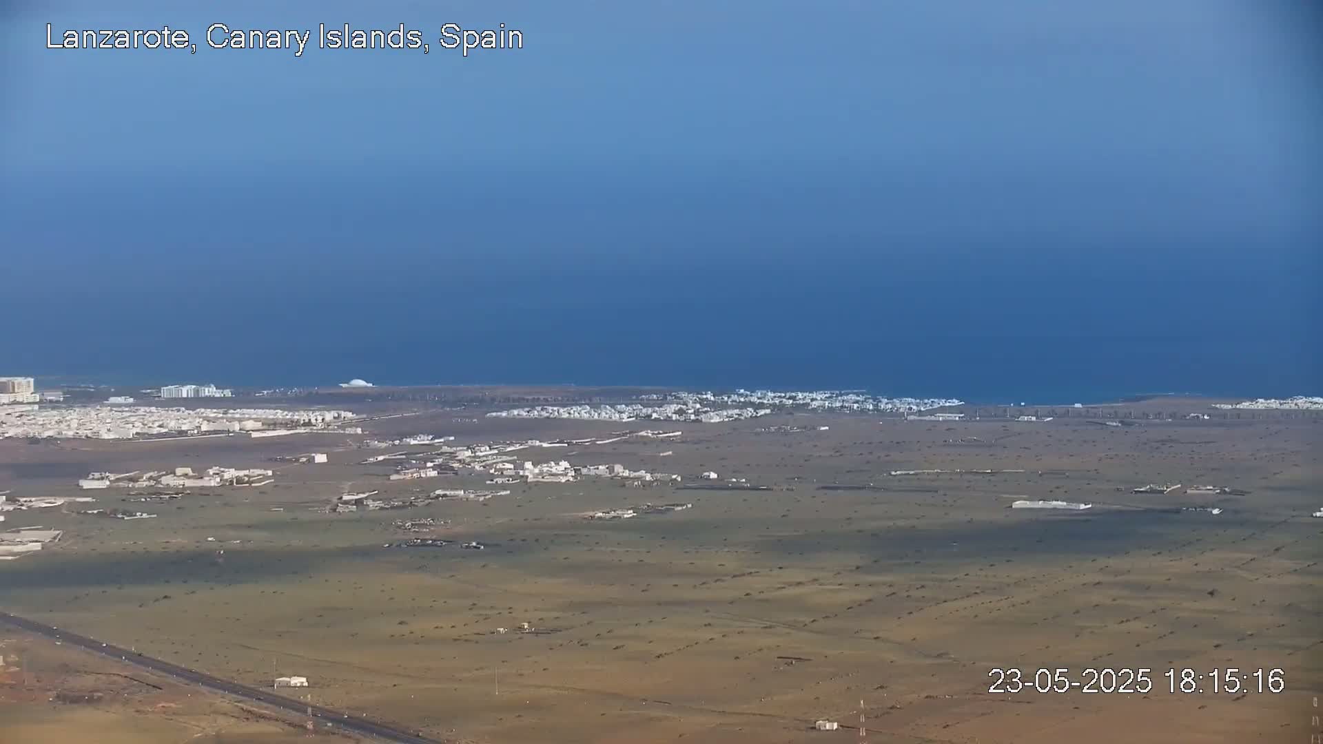 A high-angle view shows a coastal town with low-lying buildings spread across a dry, brown landscape under a clear, blue sky.