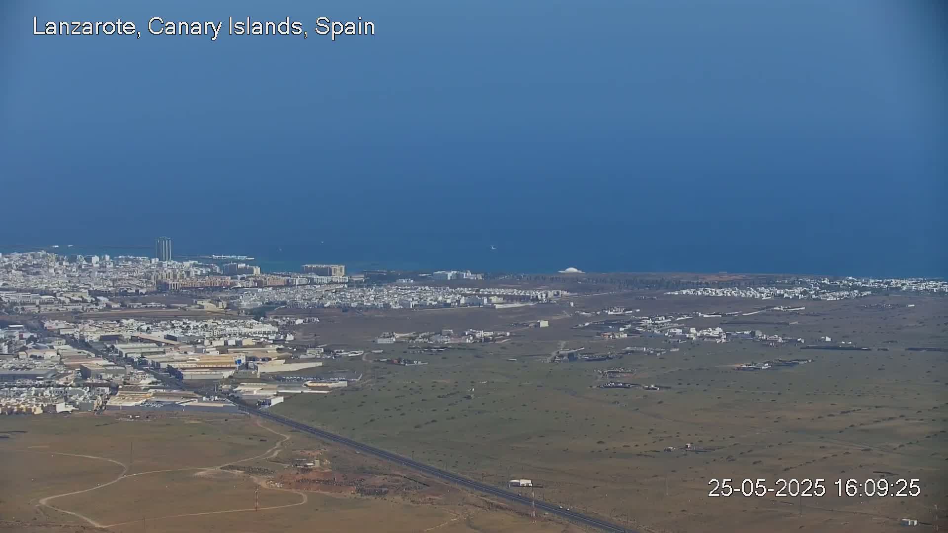 A coastal city with low-rise buildings stretches to a calm ocean under a clear blue sky.