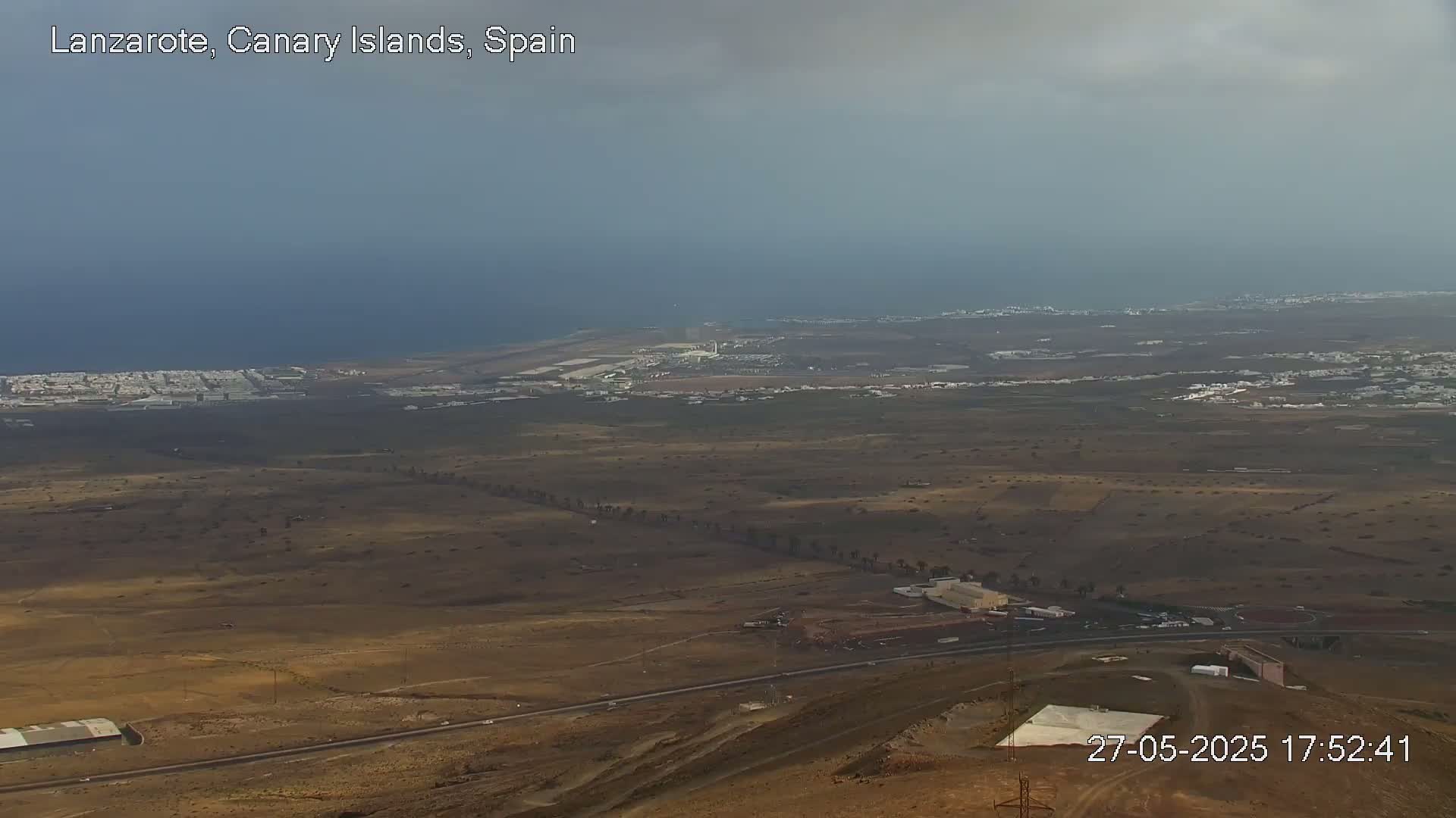 A high-angle, long shot shows a coastal area with dry, brown land, a distant ocean, and an airport under an overcast sky.