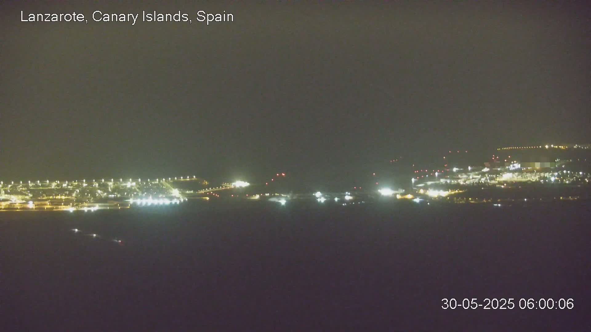 A nighttime long shot shows a brightly lit airport or industrial area under a dark, overcast sky.