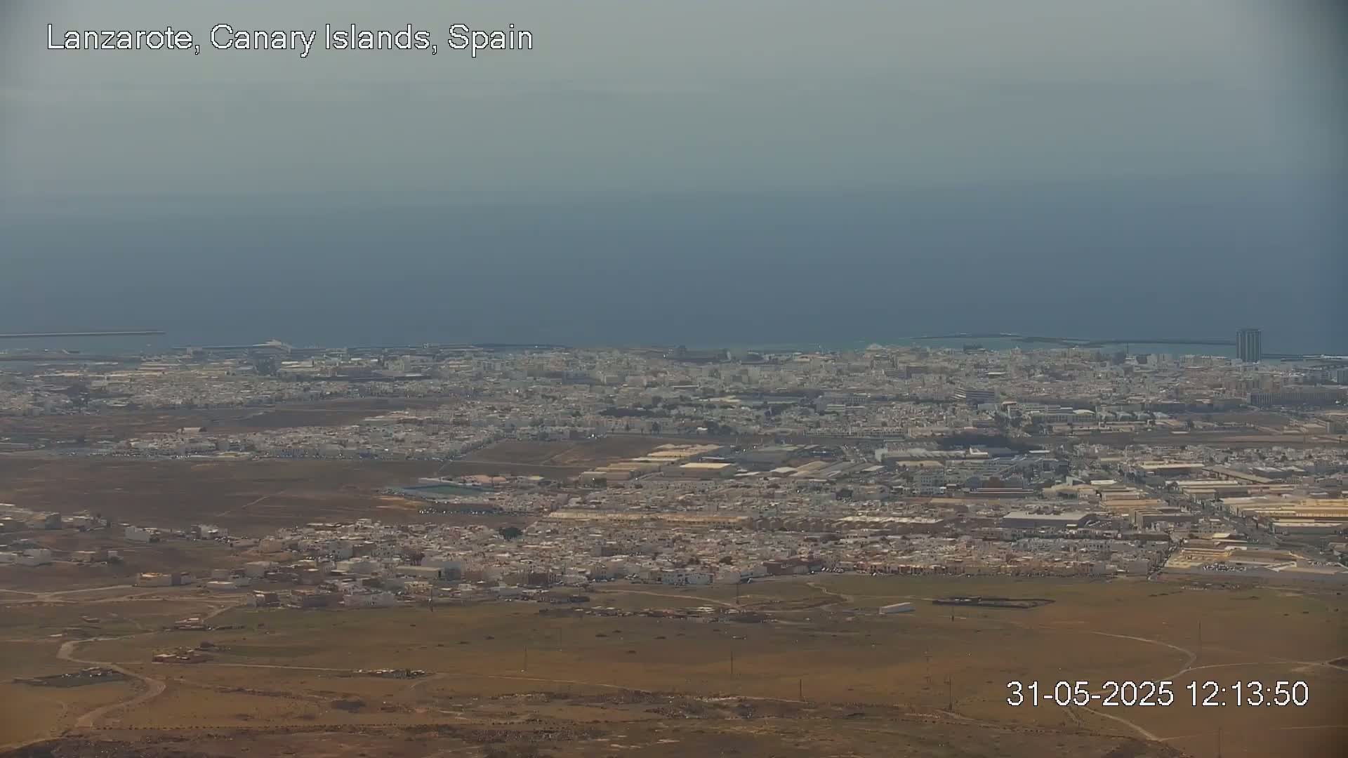 A hazy, long-distance view shows a city sprawling across a coastal plain, with a calm ocean visible in the background.