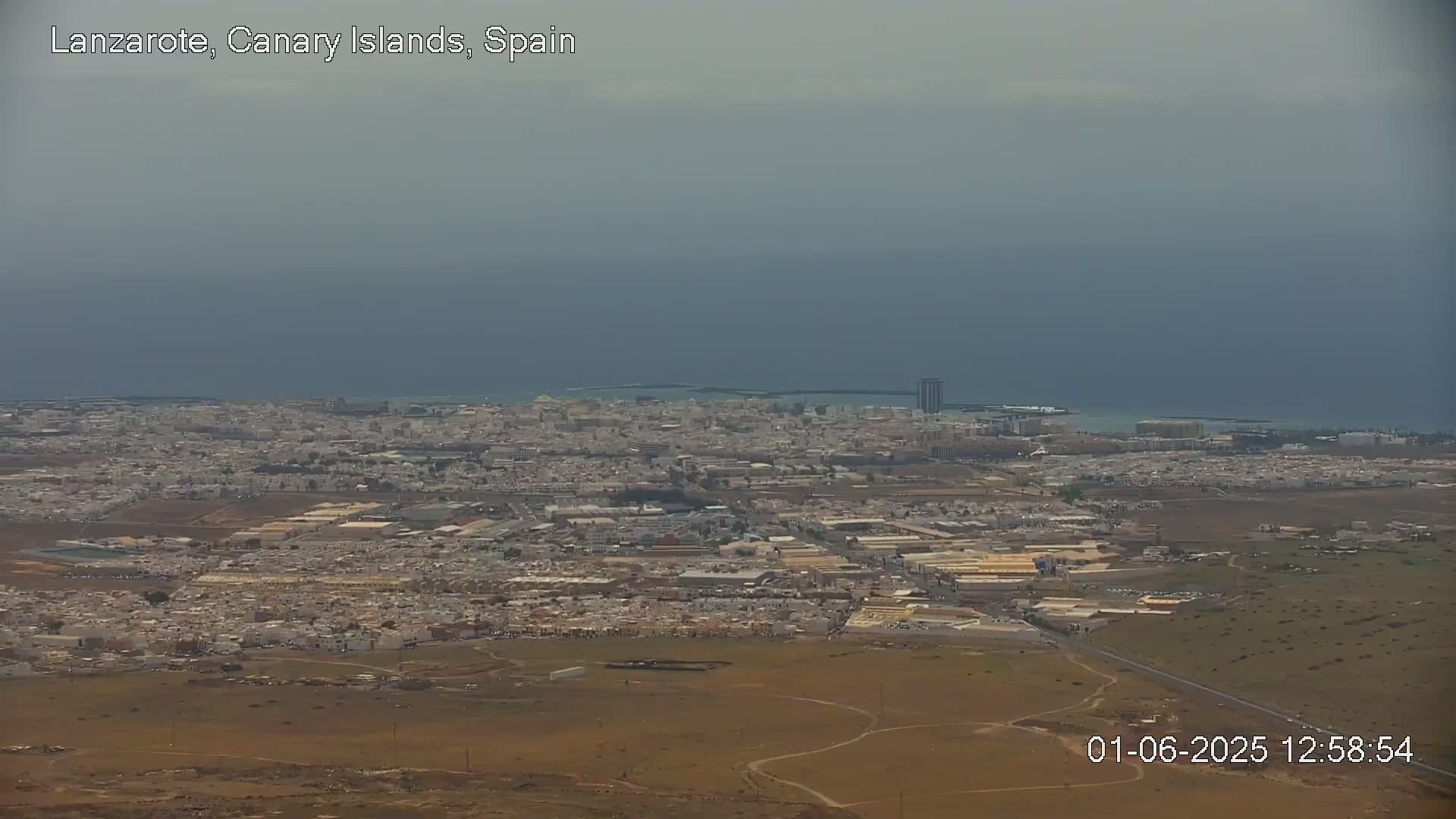 A hazy, aerial view shows a coastal city sprawling across a flat landscape, with a calm ocean in the background.