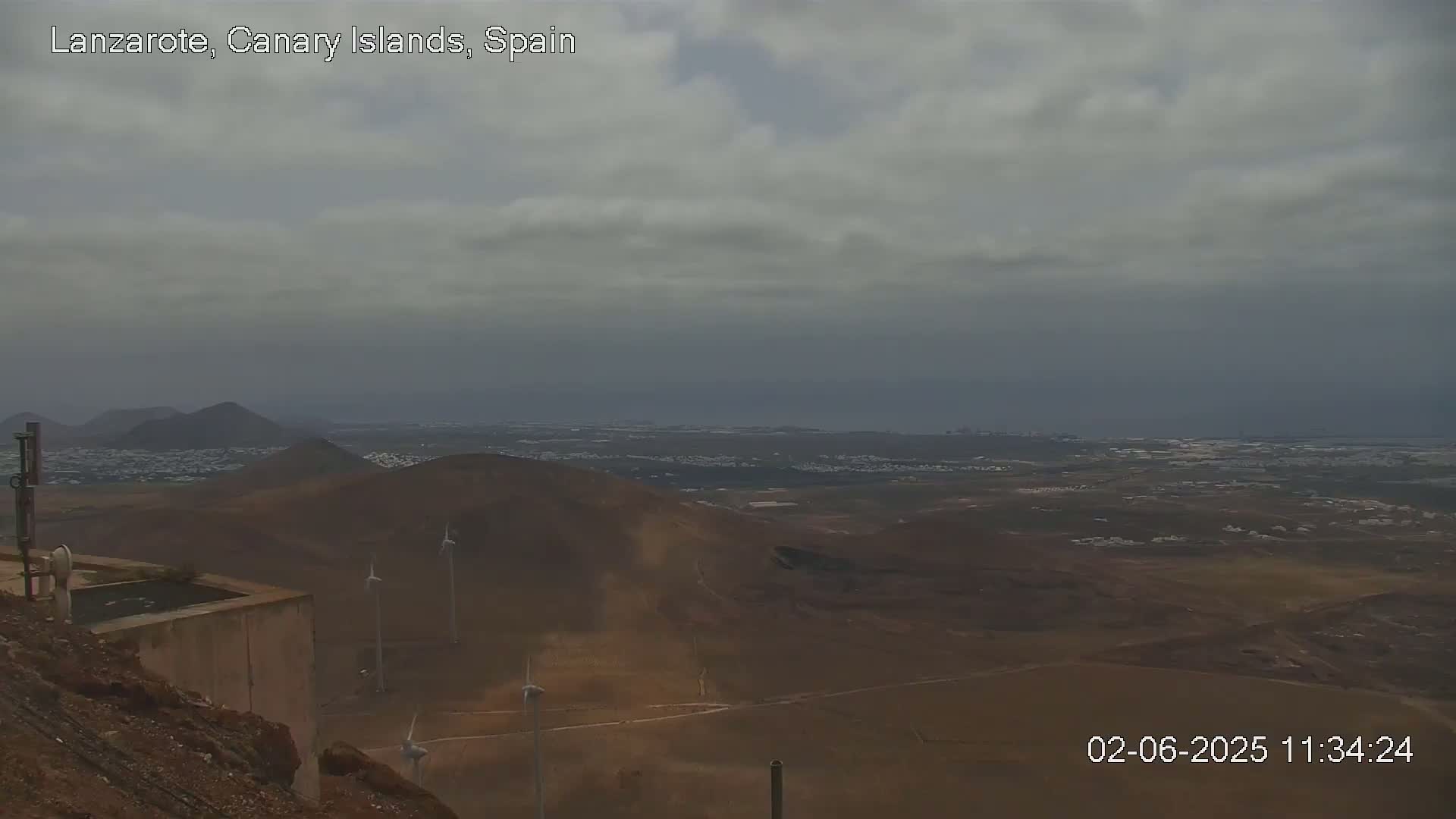 A mostly cloudy sky overlooks a brown, volcanic landscape with wind turbines and a distant ocean under a gray sky.