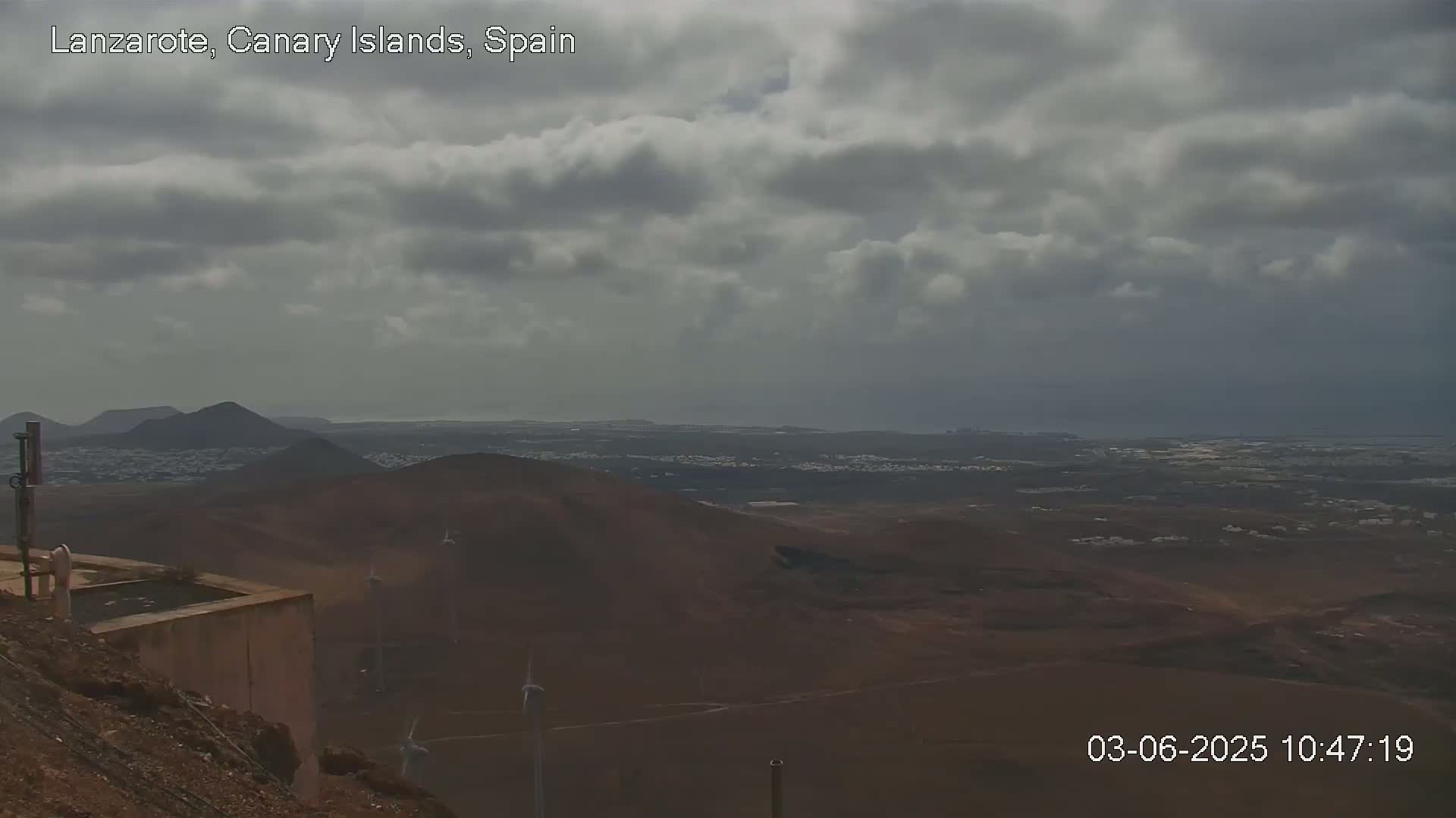 A high-angle, long shot shows a landscape of dark brown, volcanic hills under a mostly cloudy sky.