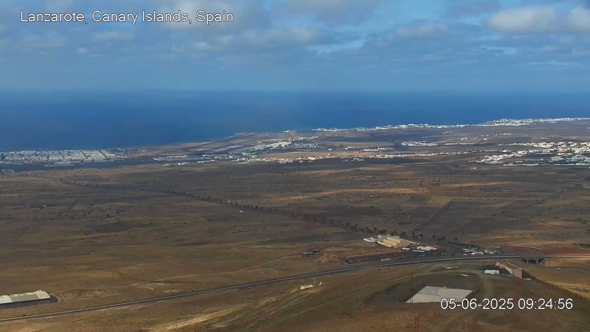 A high-angle, long shot shows a mostly arid landscape with a coastal town and ocean in the distance under a partly cloudy sky.