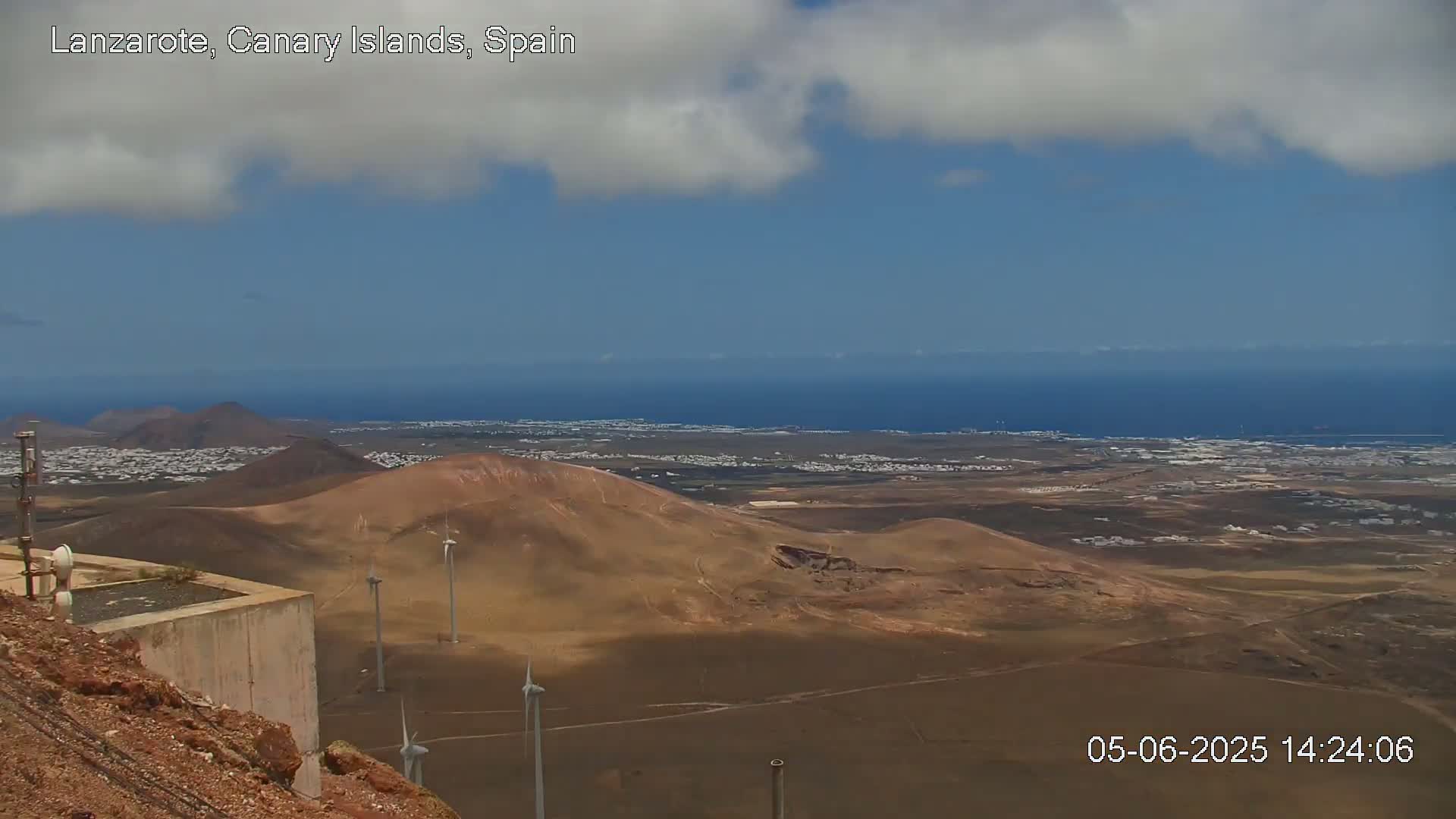 A partly cloudy day reveals a panoramic view of a volcanic landscape with wind turbines, a coastal town, and the ocean beyond.