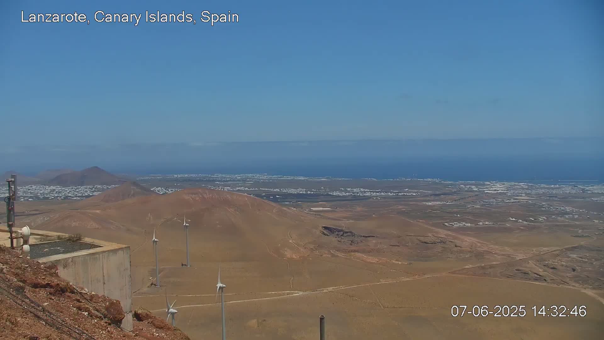 A high-angle, long shot on a clear day shows a vast, arid landscape with several wind turbines, a distant ocean, and a town nestled in the middle ground.