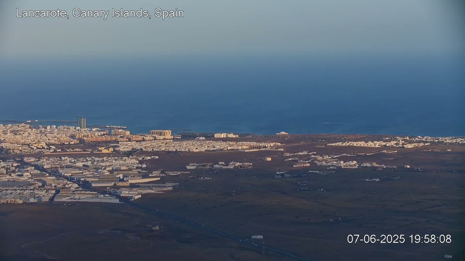 An aerial view shows a coastal town with light-colored buildings stretching towards a calm, dark blue ocean under a clear, dusky sky.