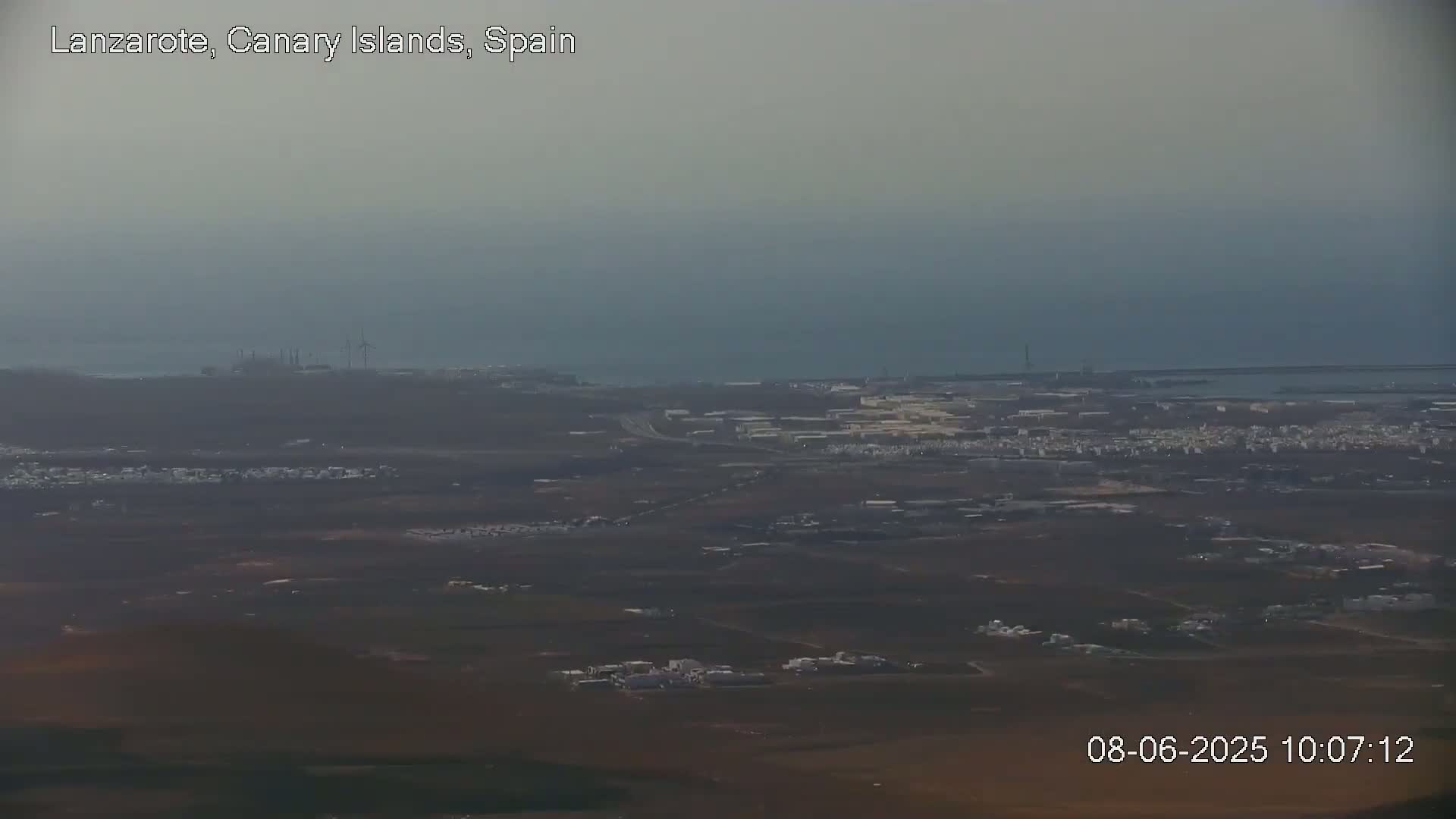 A hazy view shows a coastal town with industrial areas and wind turbines, extending to a calm ocean under a muted sky.