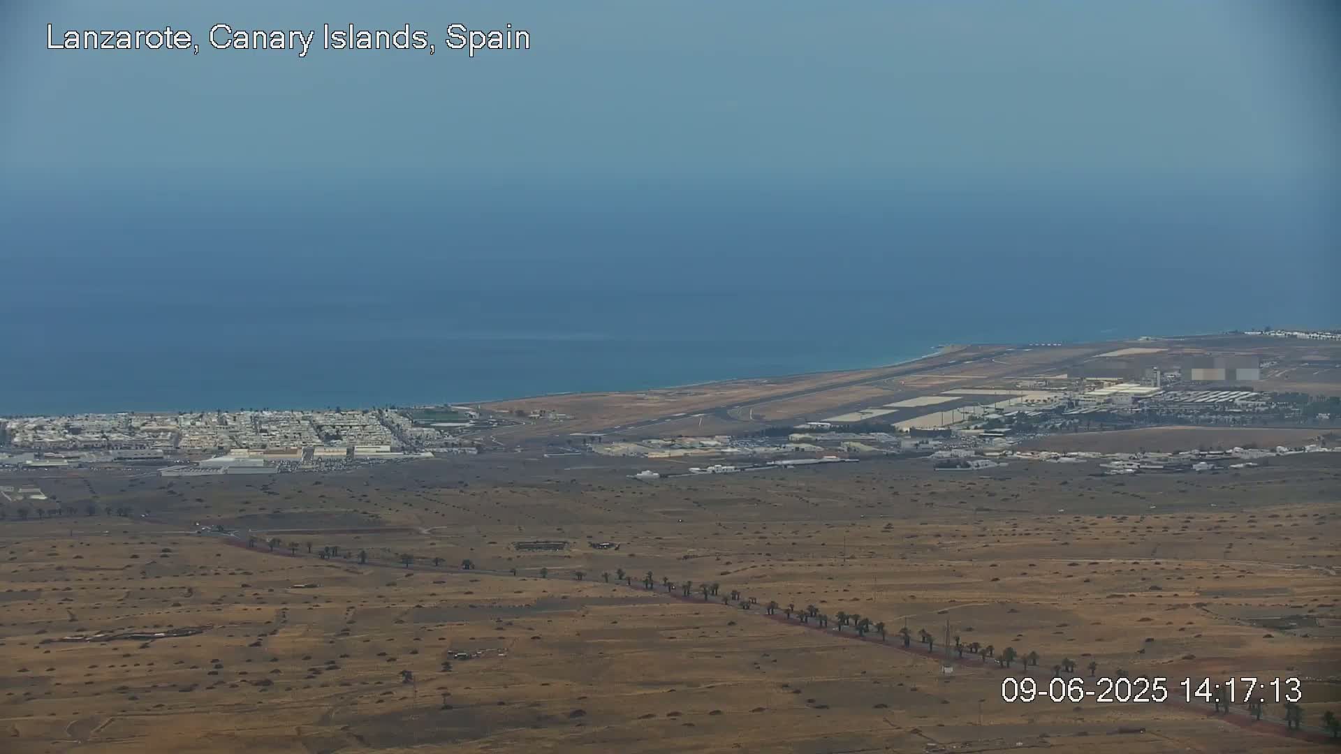 A high-angle, long shot shows a coastal airport, adjacent town, and a large expanse of arid, sparsely vegetated land under a clear, hazy sky.