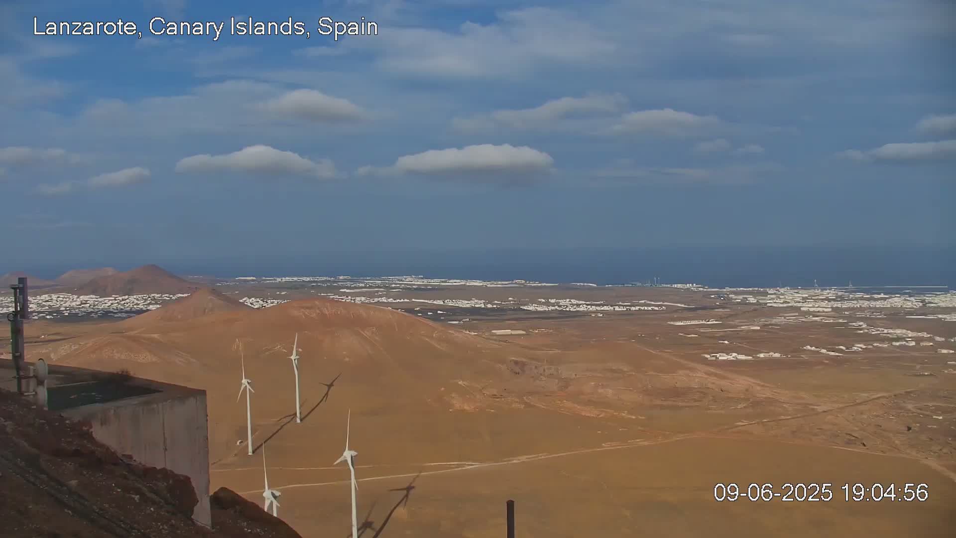 A partly cloudy sky overlooks a coastal desert landscape with several wind turbines and a distant town under a mostly clear sky.