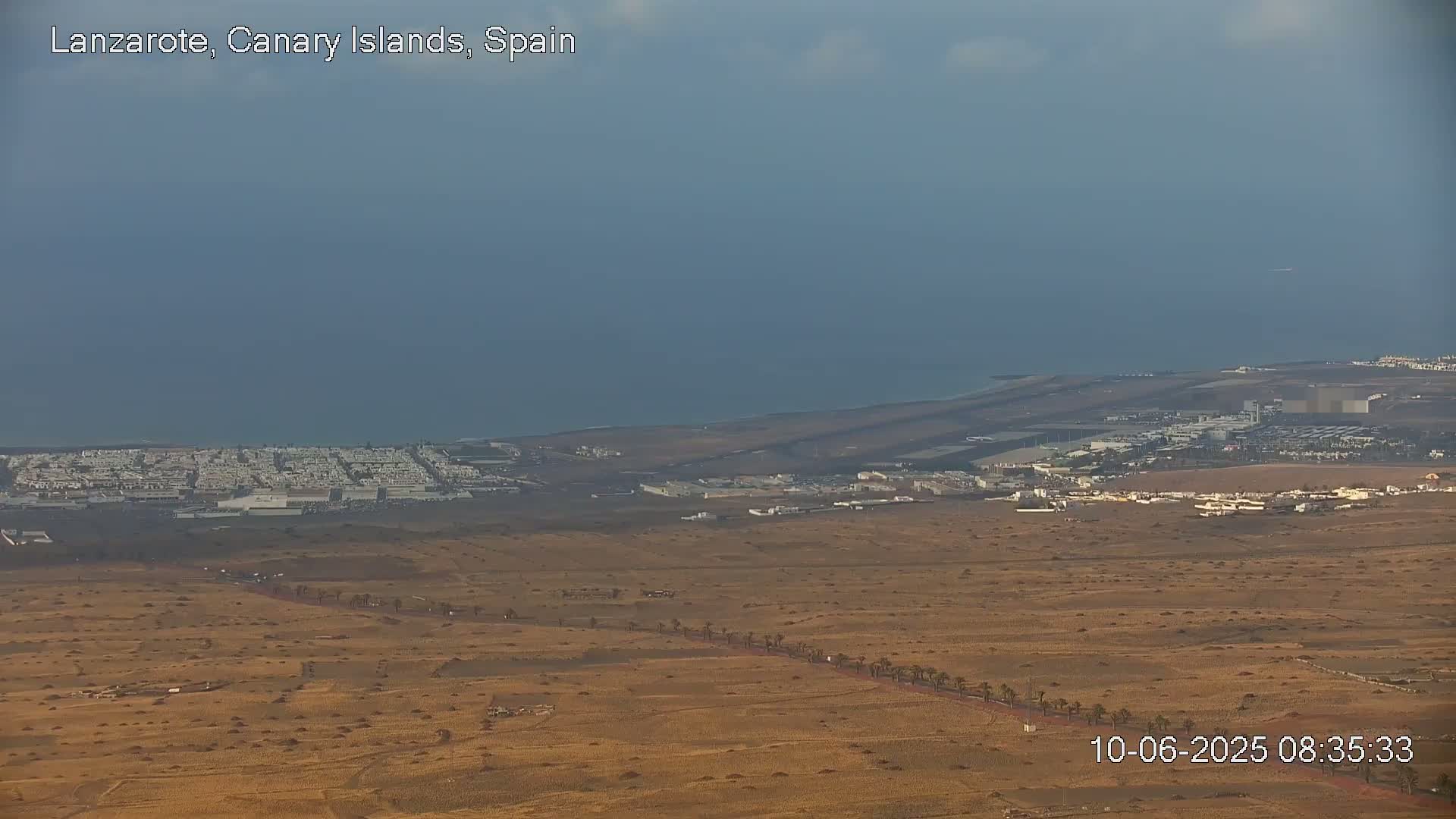 A coastal landscape under a mostly cloudy sky shows an airport and residential areas near the ocean, with a long road lined with palm trees cutting through the arid terrain.