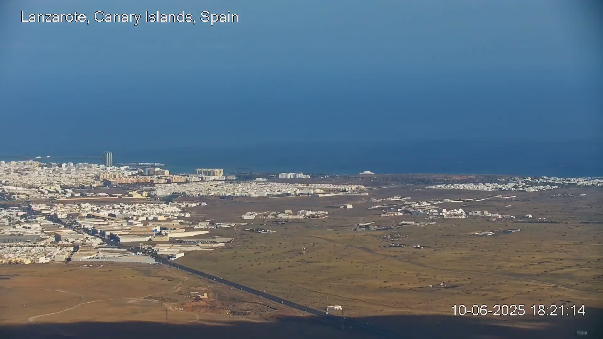 A coastal city with white buildings sprawls across a flat, arid landscape, under a clear blue sky, next to a calm ocean.