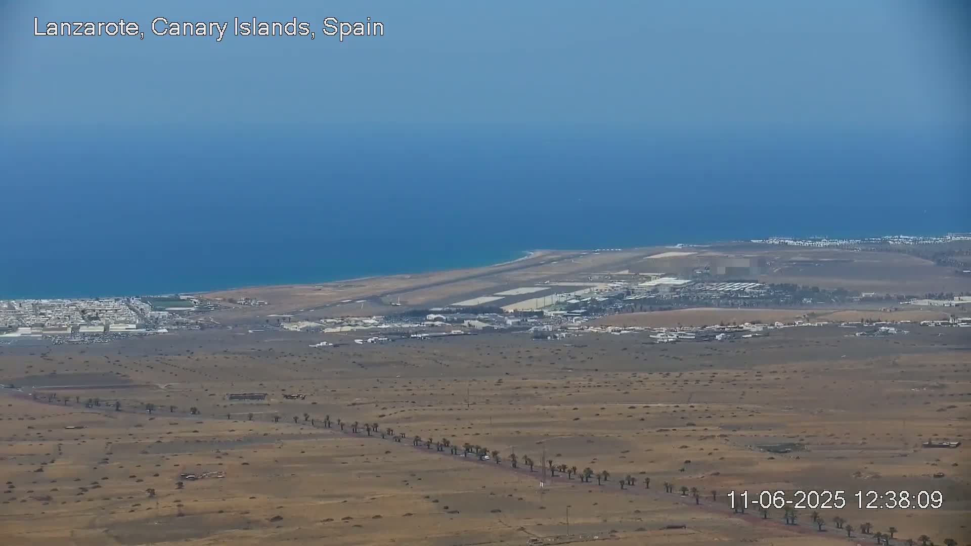 A coastal airport and surrounding arid landscape are visible under a clear sky, with a calm ocean in the background.