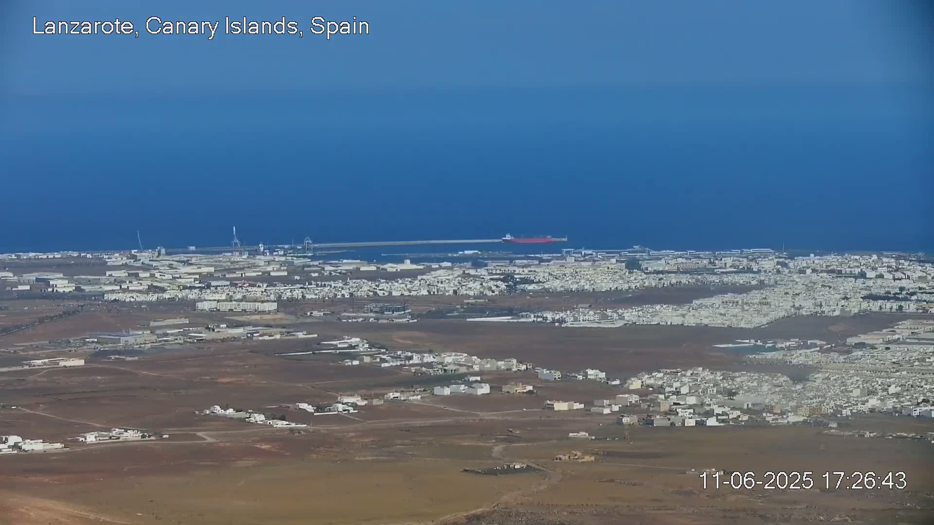 A coastal town with light-colored buildings sprawls across a dry, brown landscape under a clear blue sky, with a calm ocean and a large cargo ship visible in the distance.