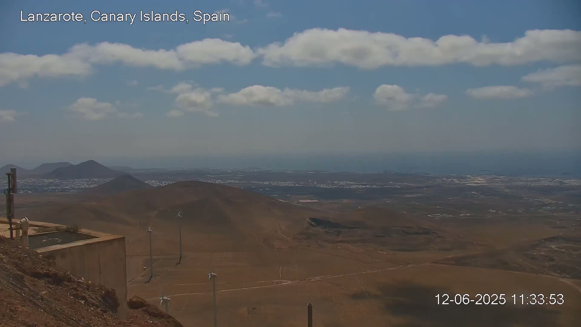 A partly cloudy, sunny day reveals a panoramic view of a volcanic landscape with a distant ocean, featuring several wind turbines in the foreground.