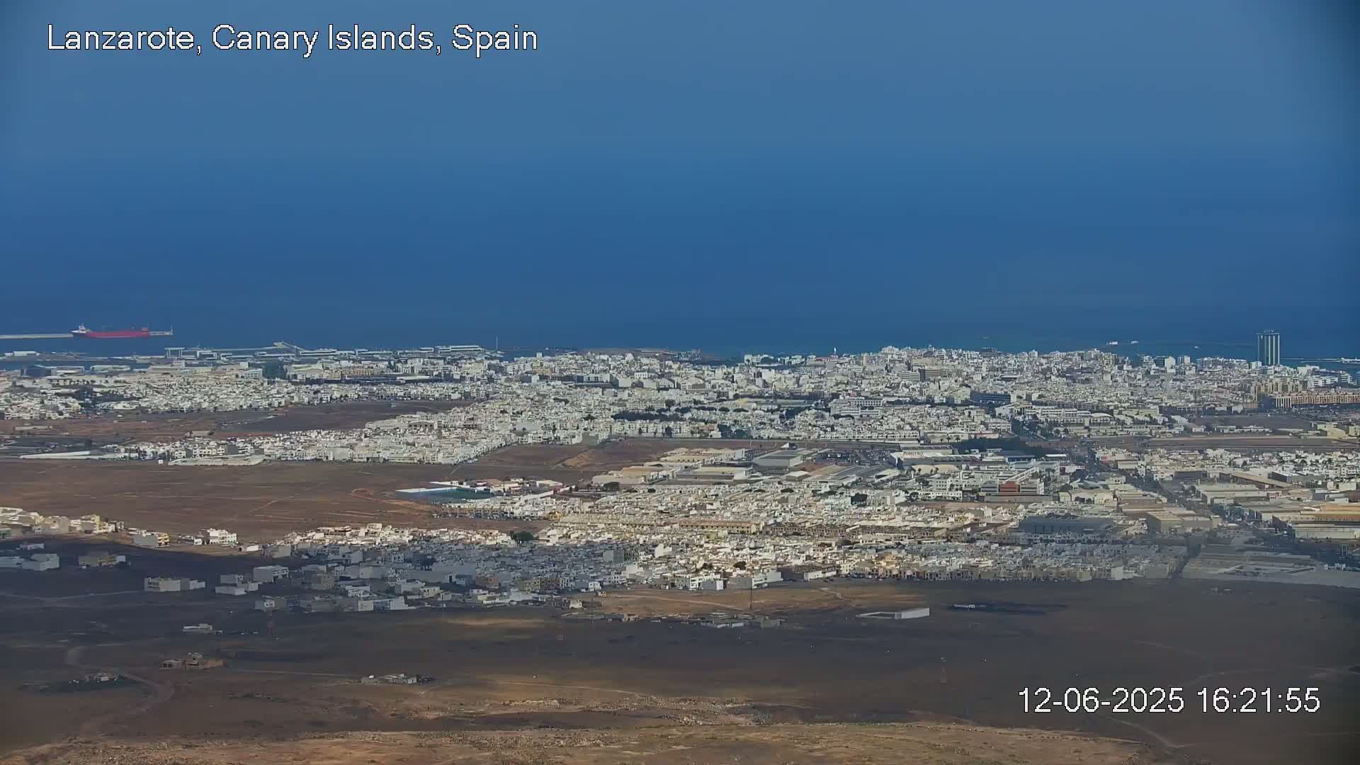 A sprawling coastal city with light-colored buildings extends to a calm blue ocean under a clear blue sky.