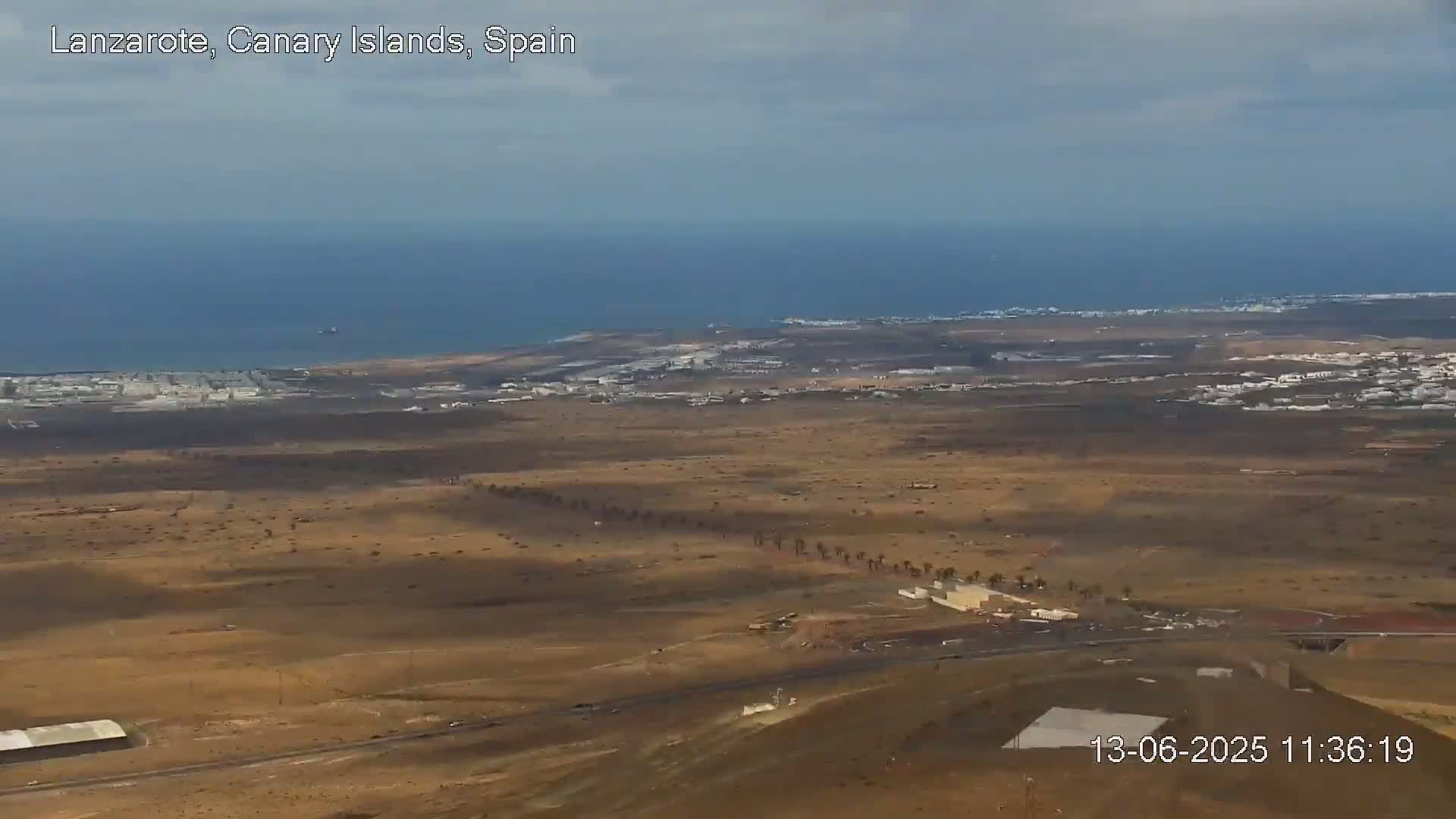A high-angle view shows a mostly-flat, arid landscape with sparse vegetation, a coastal town, and the ocean under a partly cloudy sky.