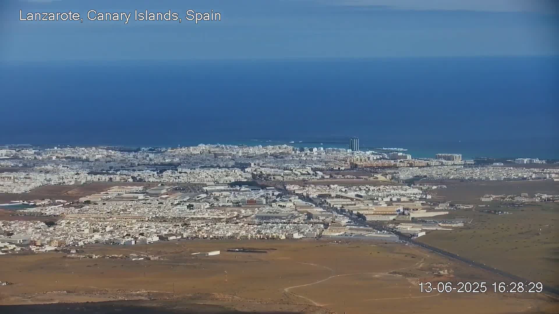 A high-angle, long shot shows a coastal city on a clear, sunny day, with a large expanse of ocean visible in the background.