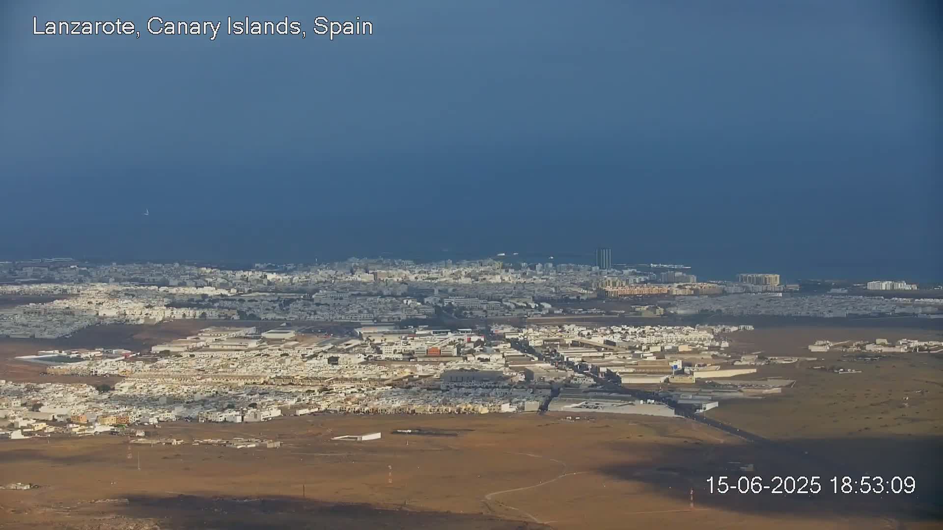 An expansive view of a coastal city with light-colored buildings, under a mostly cloudy sky, shows a dark expanse of ocean in the distance.