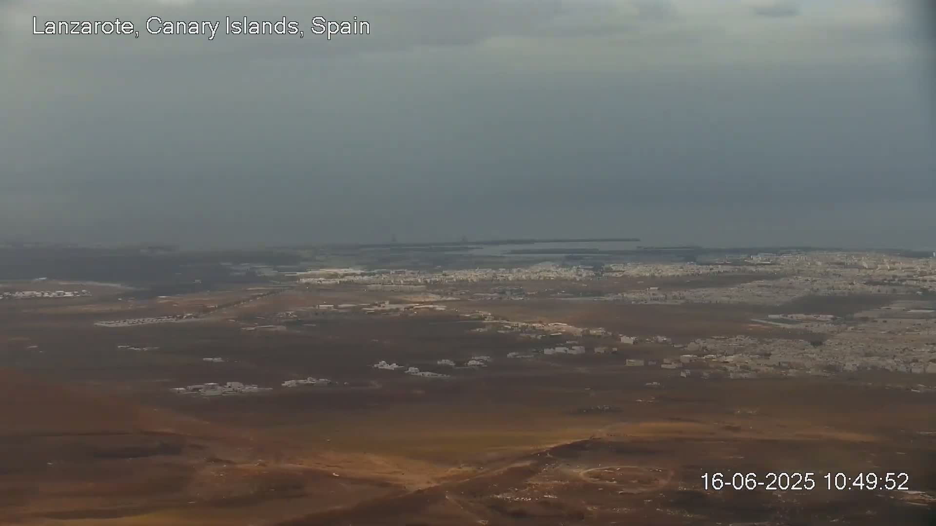 A sprawling, low-lying town with white buildings extends across a brown, arid landscape under an overcast sky, with a body of water visible in the distance.
