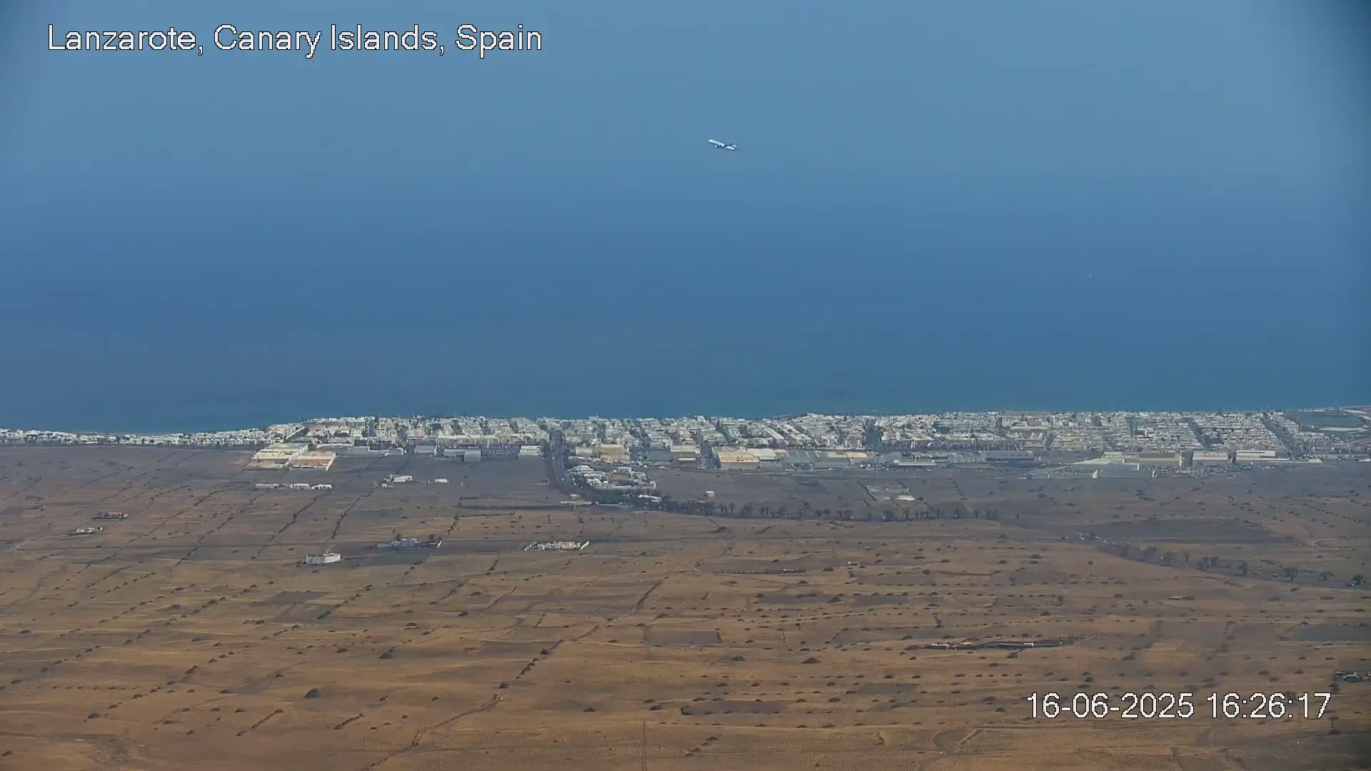 A high-altitude view shows a coastal town and arid, sparsely vegetated landscape under a clear blue sky, with an airplane visible in the distance.
