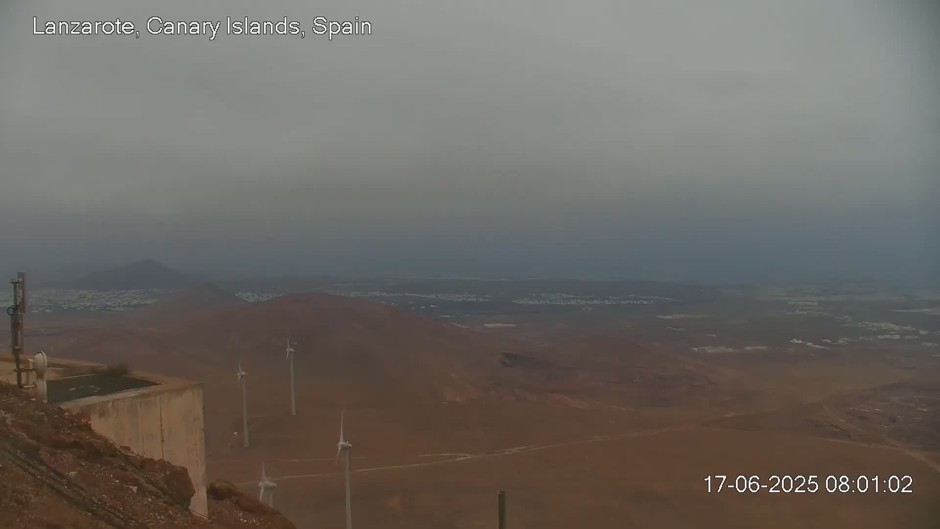 A hazy, overcast view shows a vast, arid landscape with several wind turbines and a distant town under a gray sky.