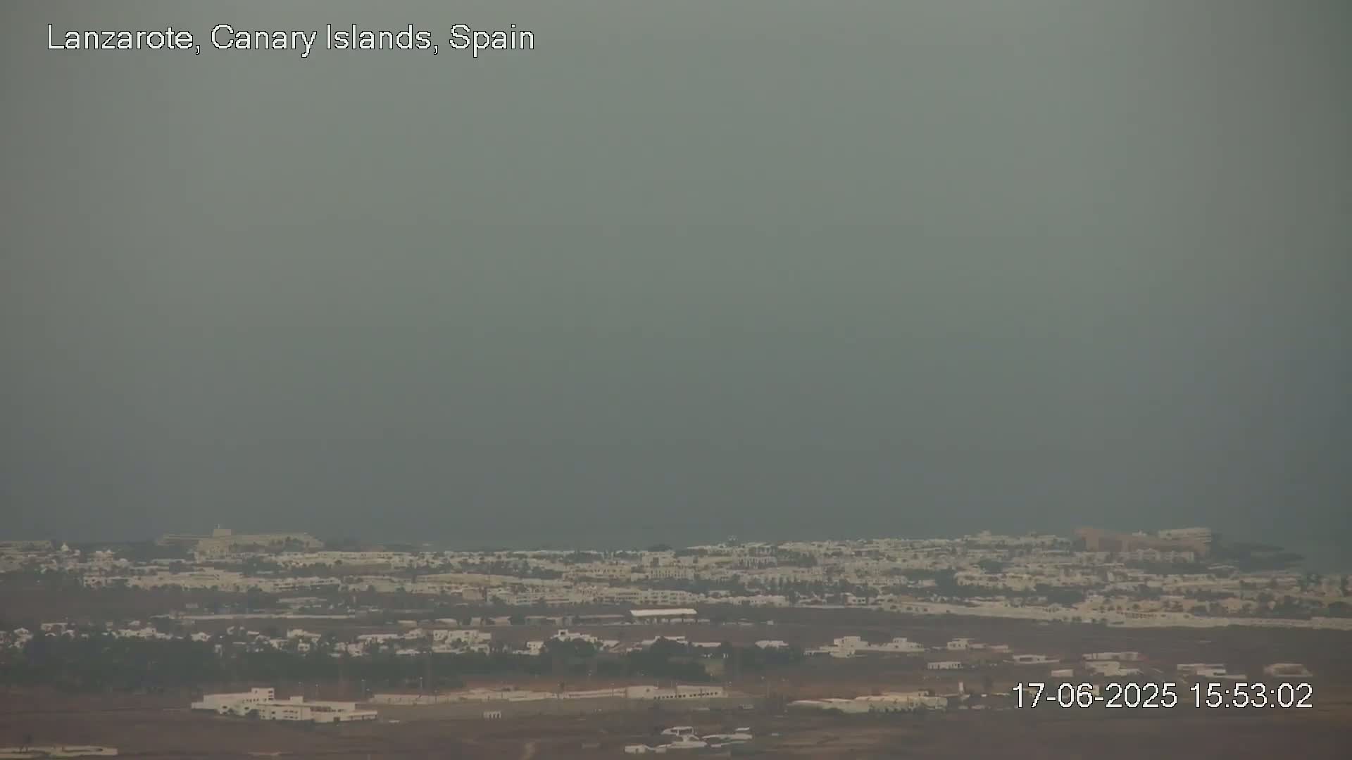 A hazy, overcast view shows a coastal town with low-lying buildings extending to a gray ocean.