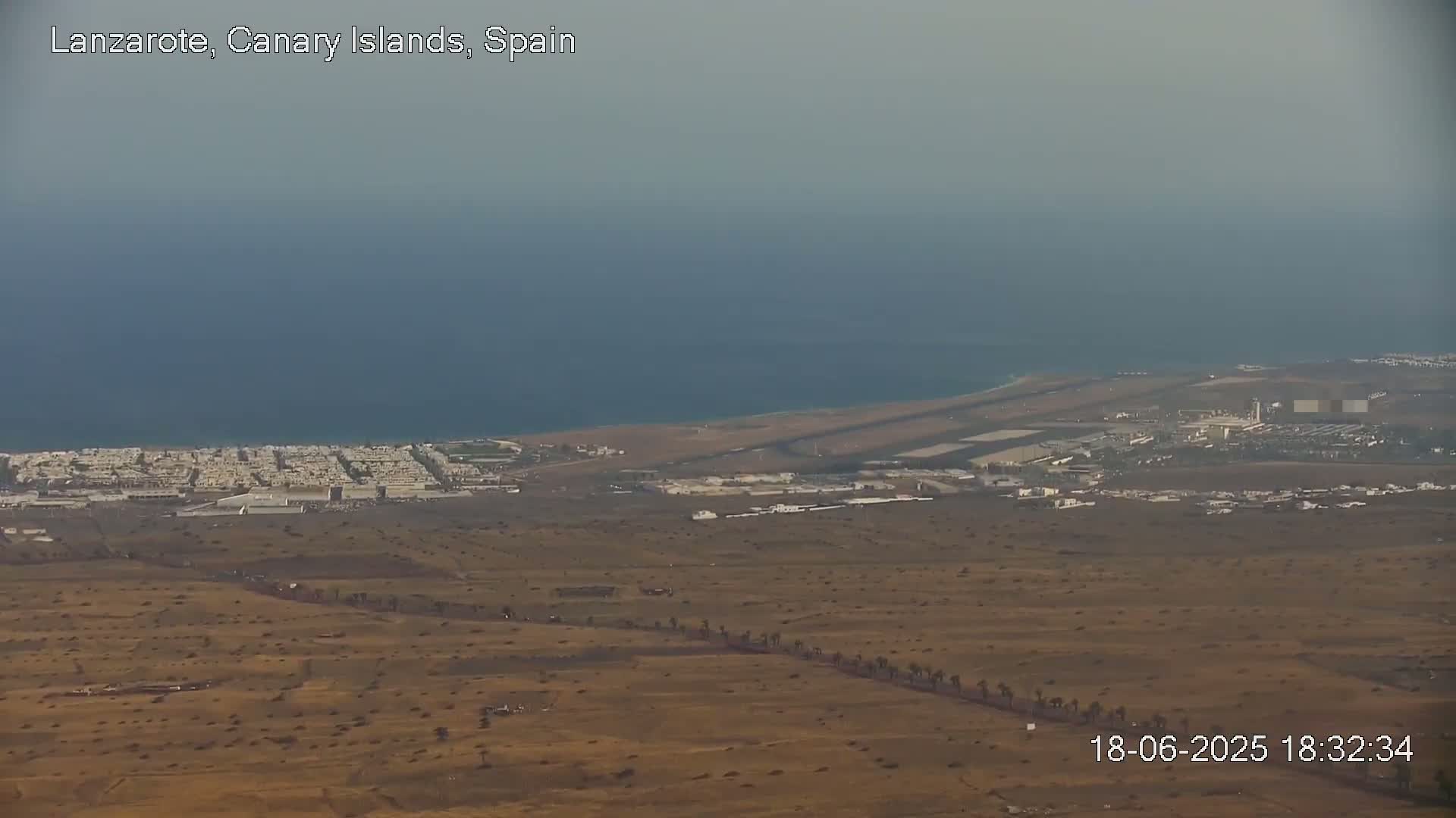 An aerial view shows a coastal airport and town in a hazy, dry landscape next to a calm ocean.