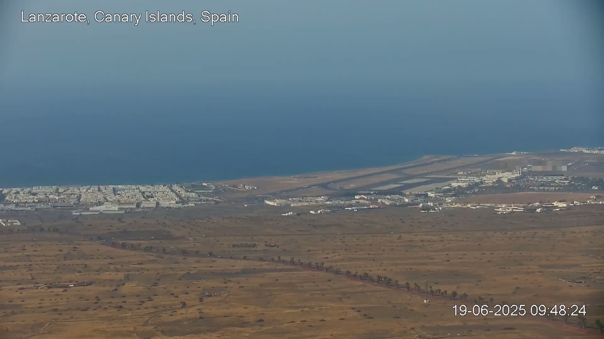 An aerial view shows a coastal airport and surrounding buildings next to a calm ocean under a hazy sky.